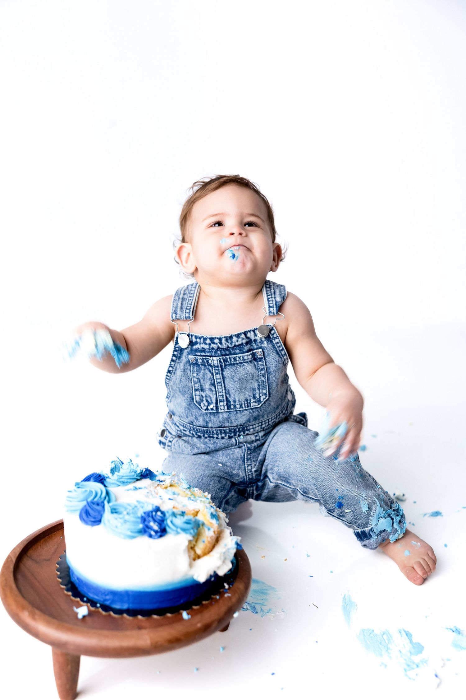 Baby in denim overalls smashes blue and white cake during first birthday photoshoot.
