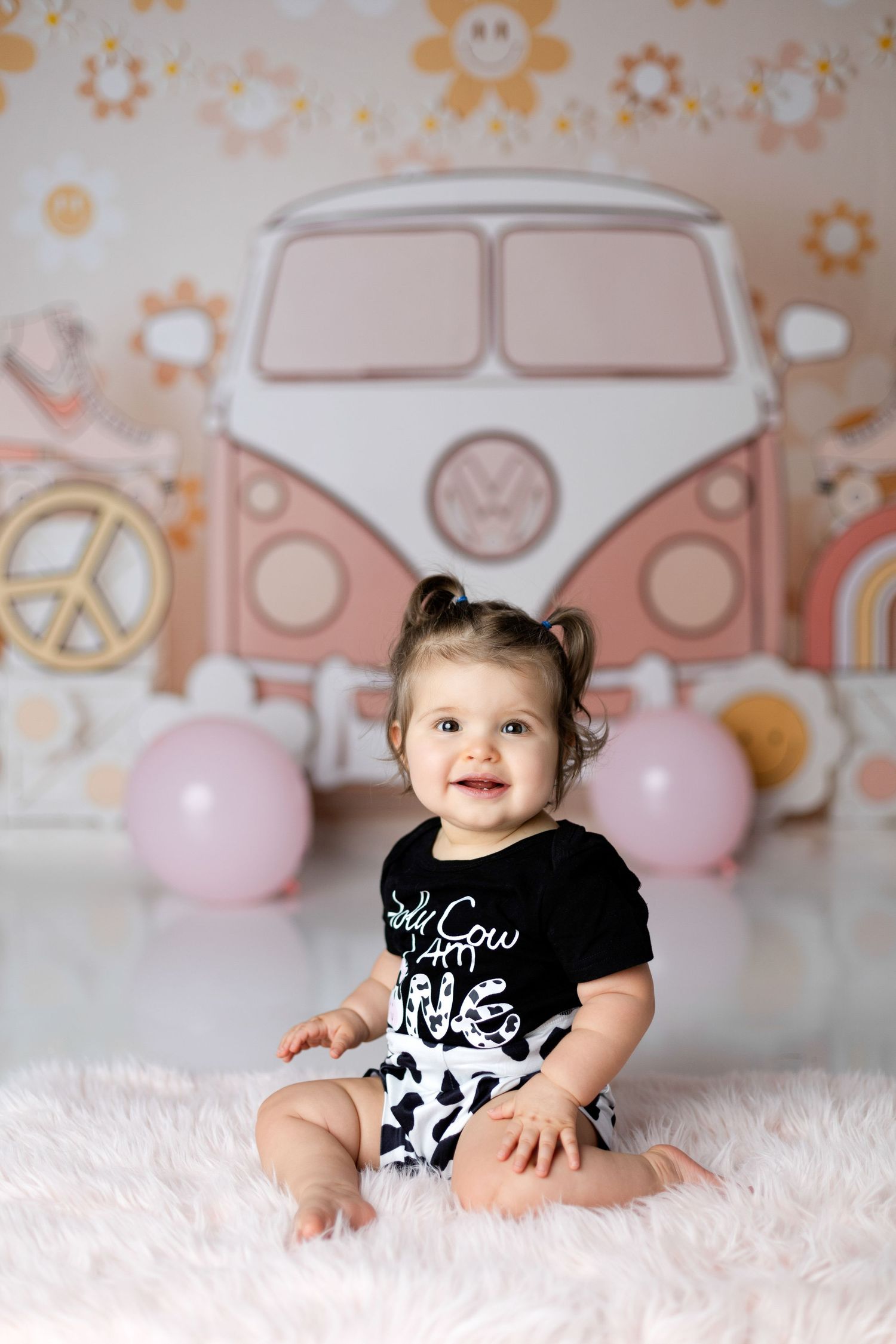 Baby in black onesie lying on white rug with retro car backdrop and pink balloons.