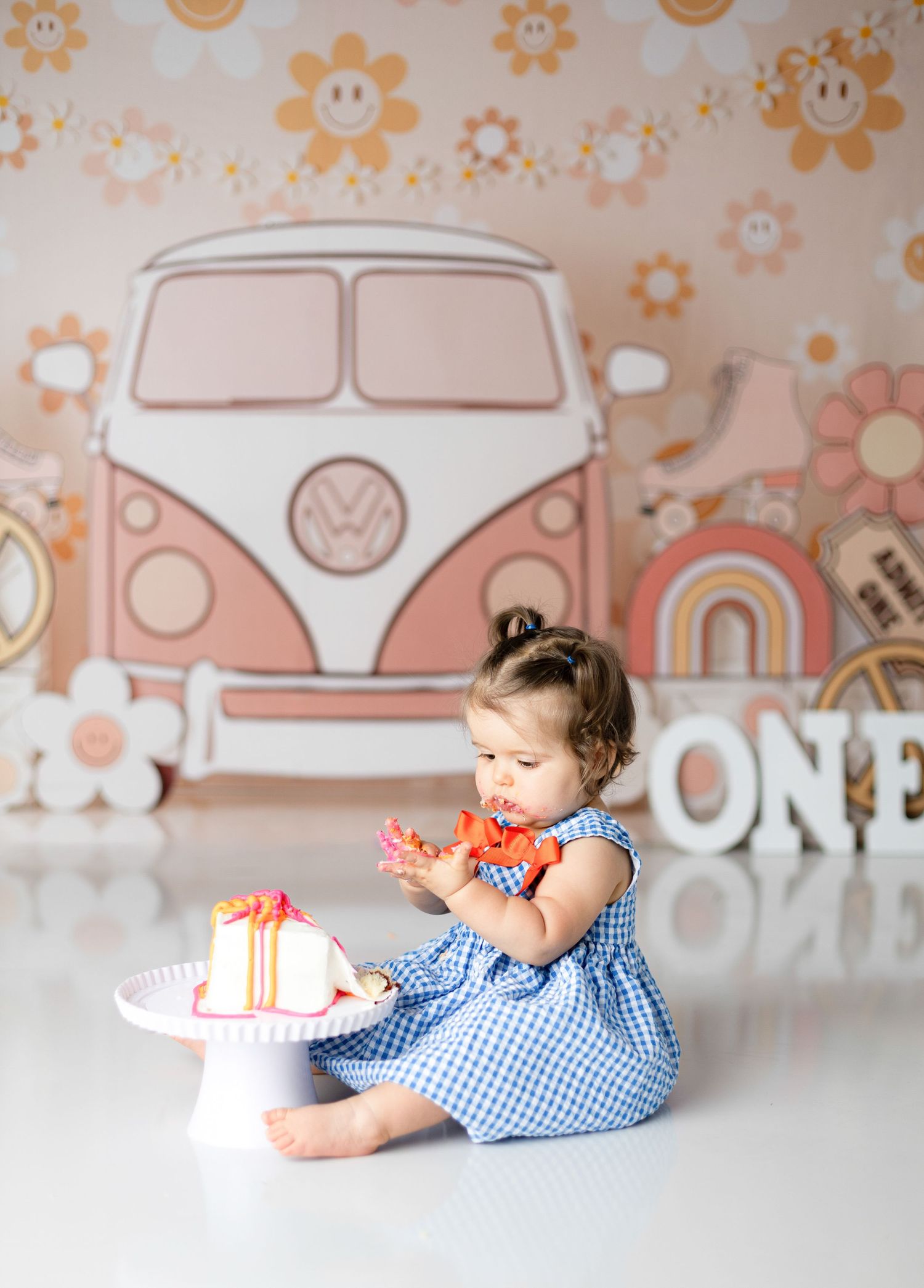 Baby in blue gingham dress reaching for birthday cake with vintage car themed backdrop.