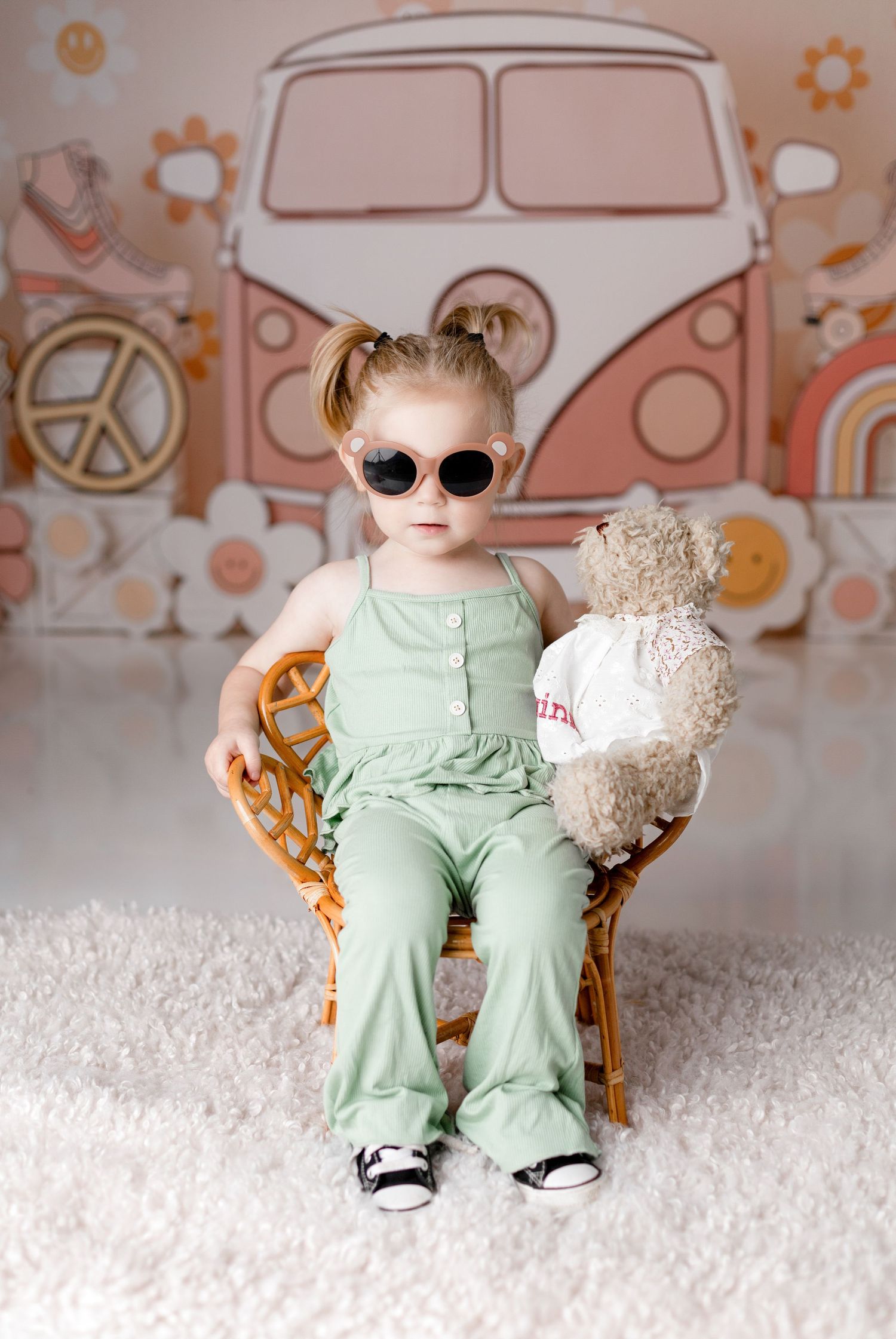 Stylish toddler wearing sunglasses poses with teddy bear against retro van backdrop.