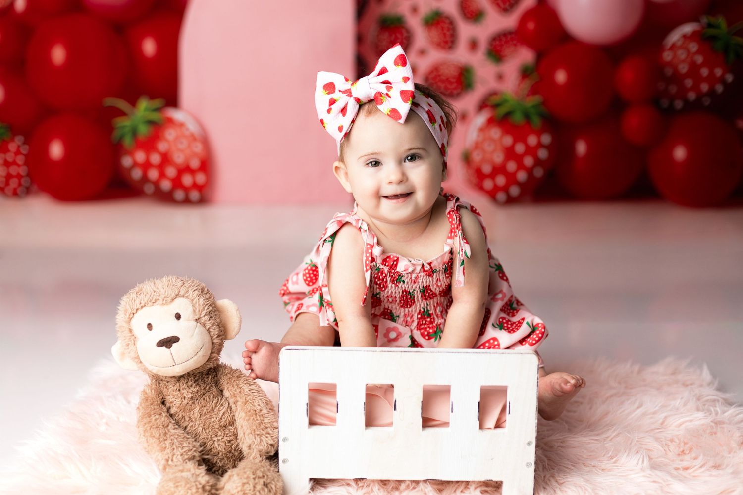 Baby sitting in white frame with red strawberry themed backdrop and balloons.