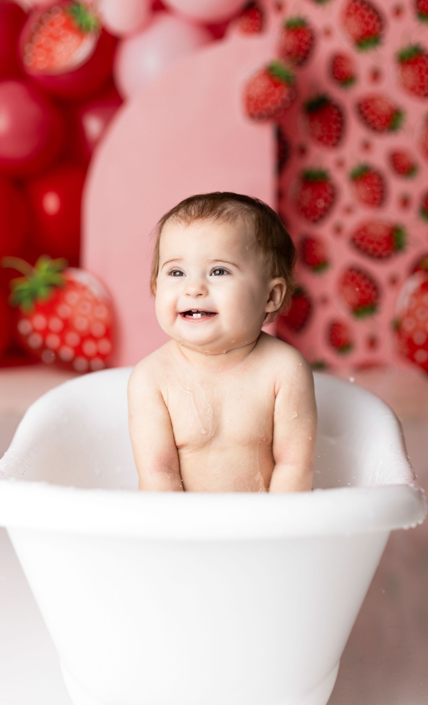 Baby sitting in white bathtub against strawberry patterned backdrop with pink balloons.