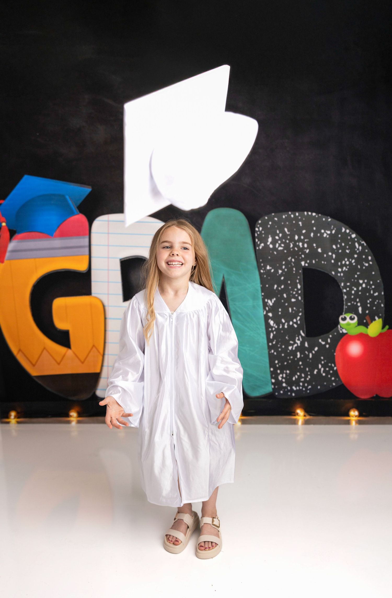 A child in a white graduation gown poses against a decorated blackboard background.