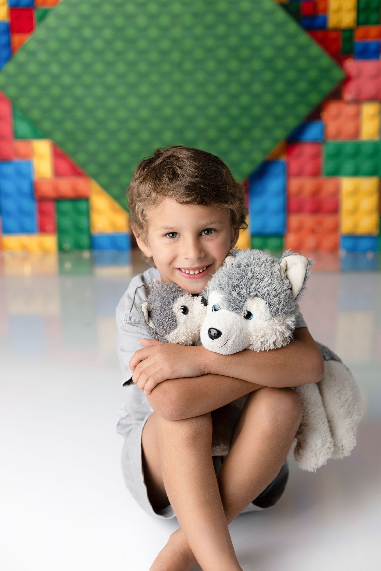 Child holding grey stuffed animal while sitting against green LEGO wall background.