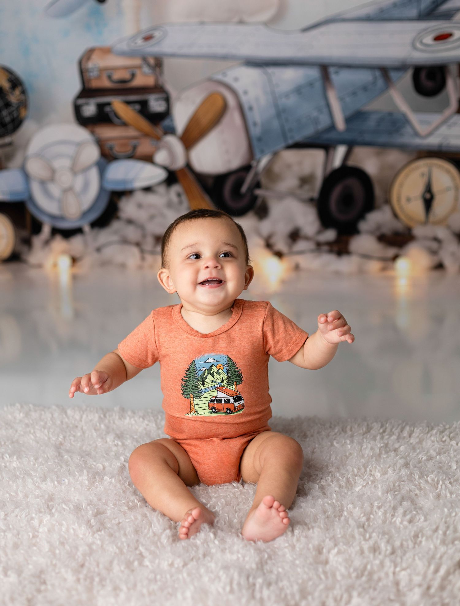 Baby in orange onesie sitting on white fluffy rug with vintage car decorations in background.