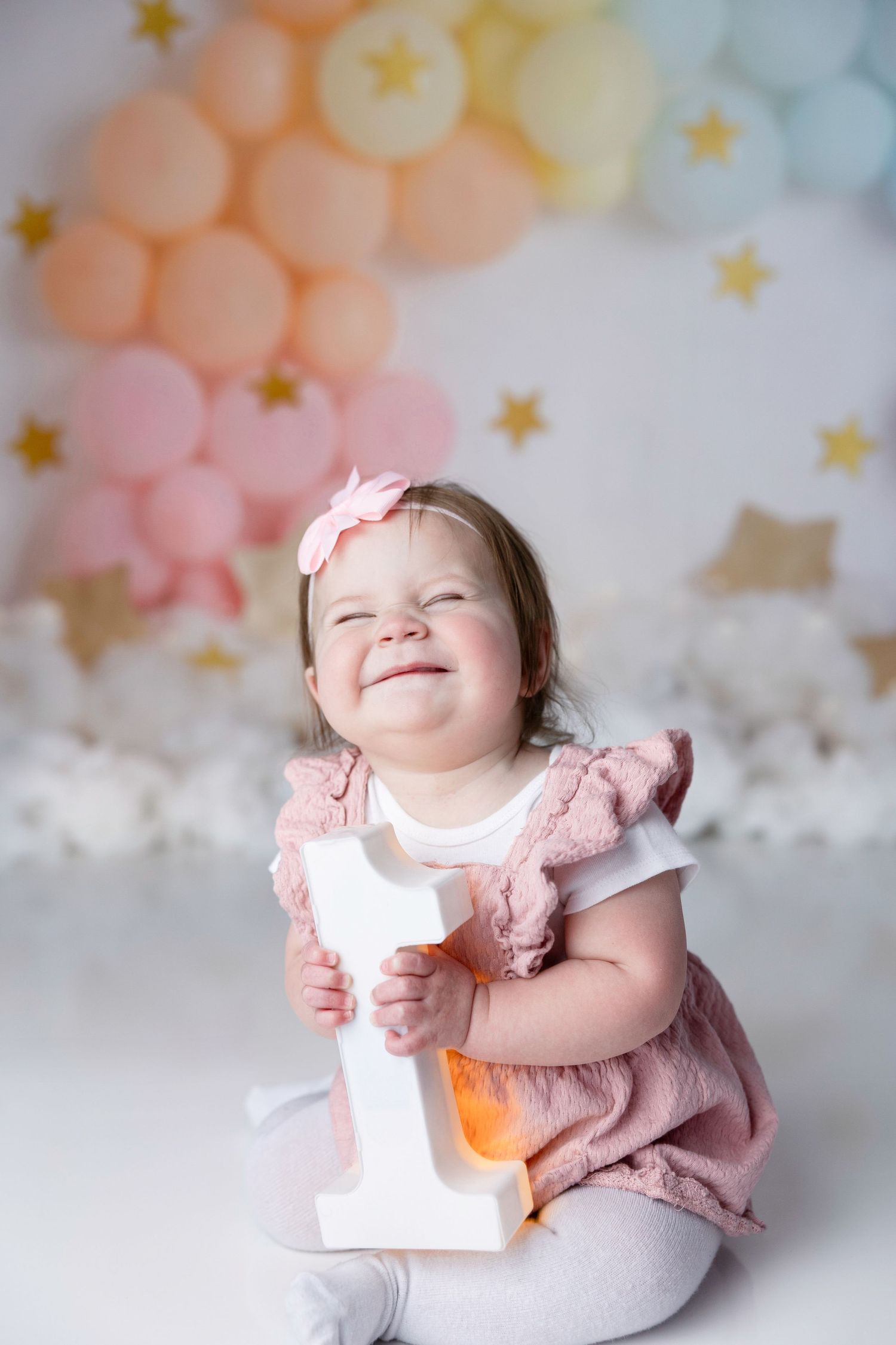 Baby in pink outfit smiling during cake smash photoshoot with pastel balloon backdrop.