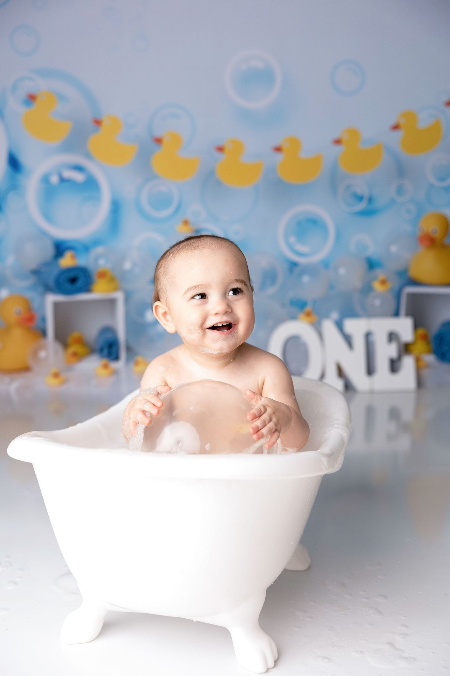 Baby bath photoshoot with rubber duck theme and blue bubble backdrop.