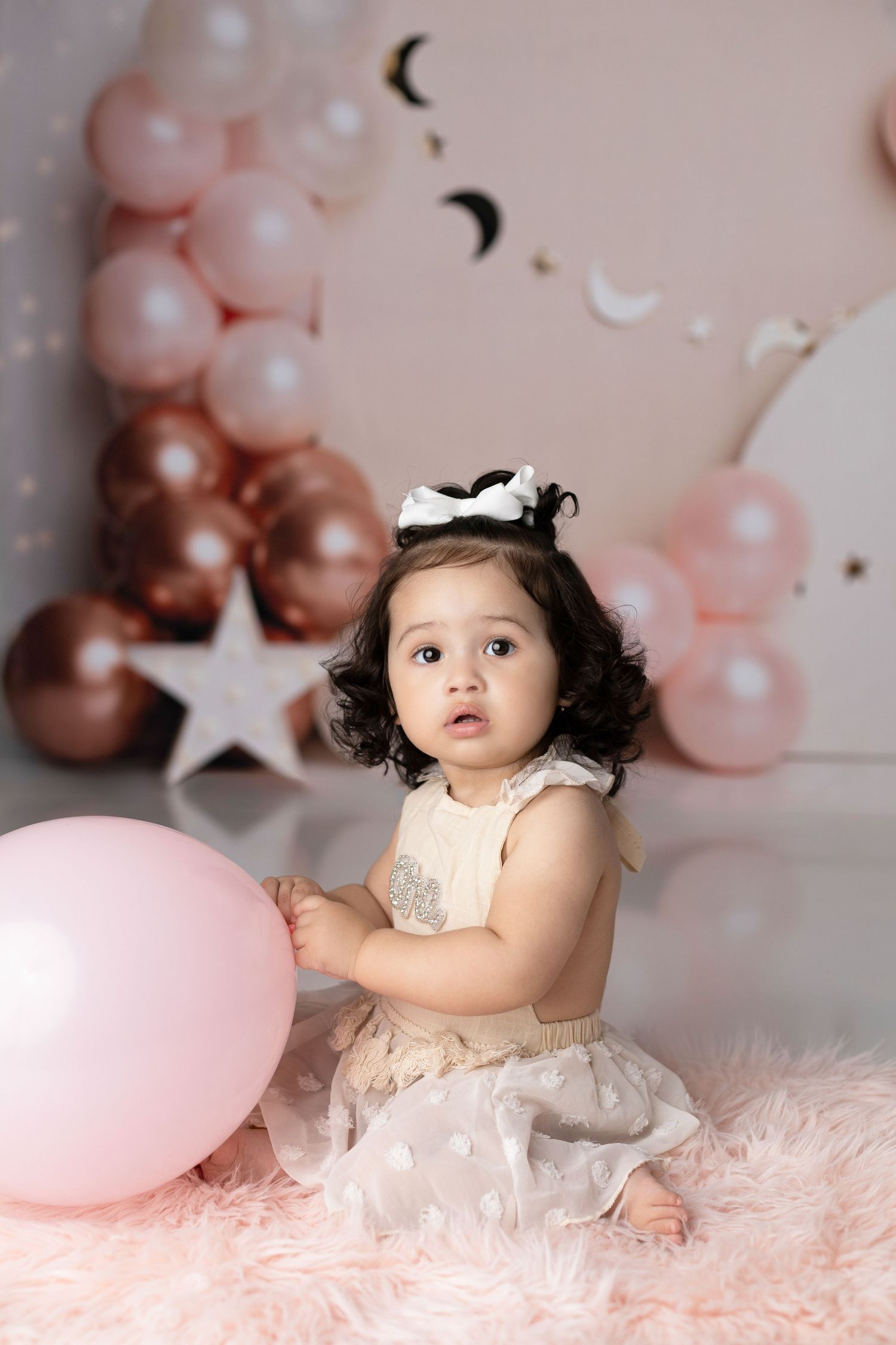 Toddler in cream dress with party decorations and pink balloons in background.