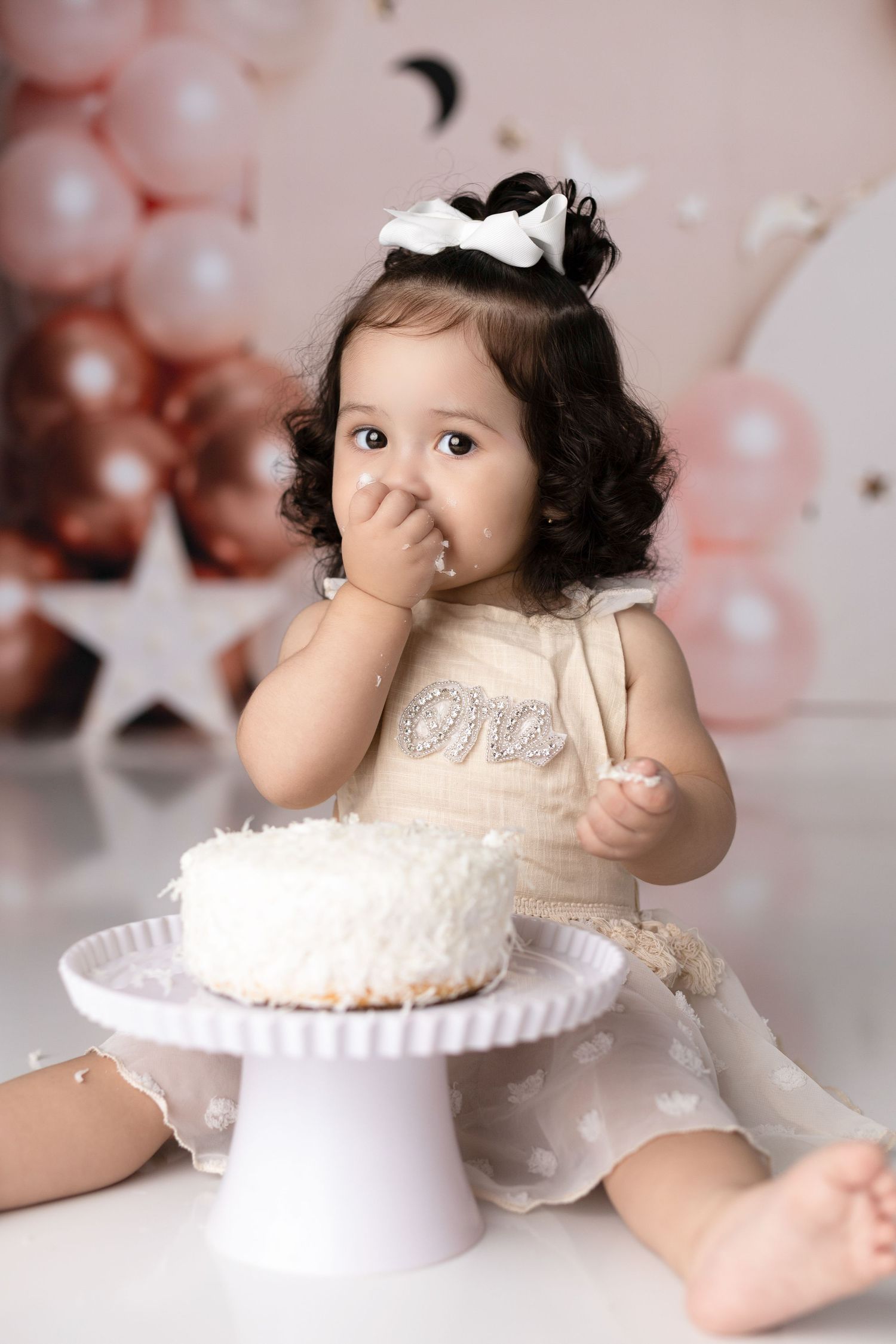 Toddler in lace dress sitting with white cake on stand and party decorations.