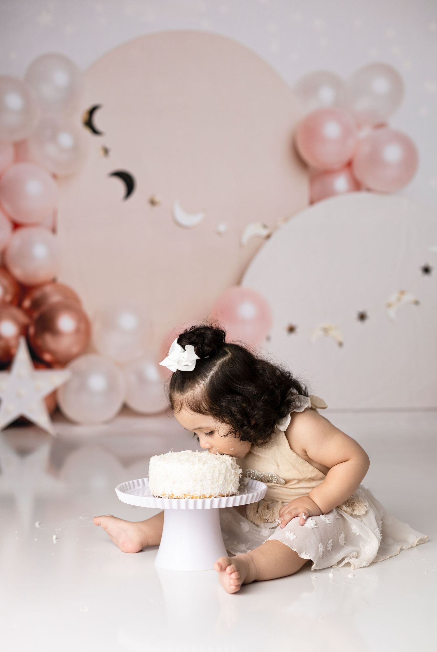 Baby cake smash photoshoot with pink balloons and white frosted cake on white backdrop.