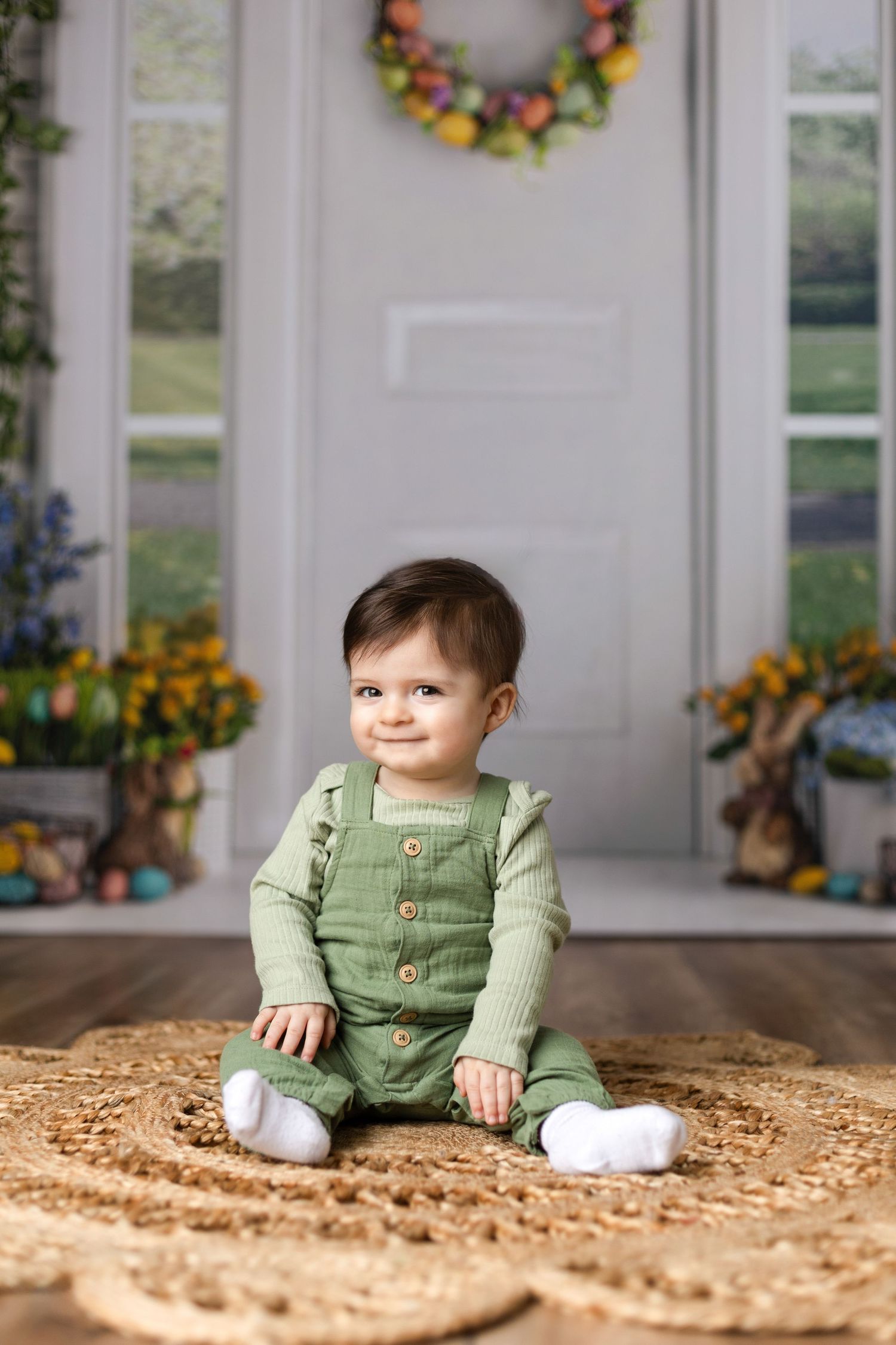 Baby in green overalls sitting on a textured brown surface near white paneled walls.
