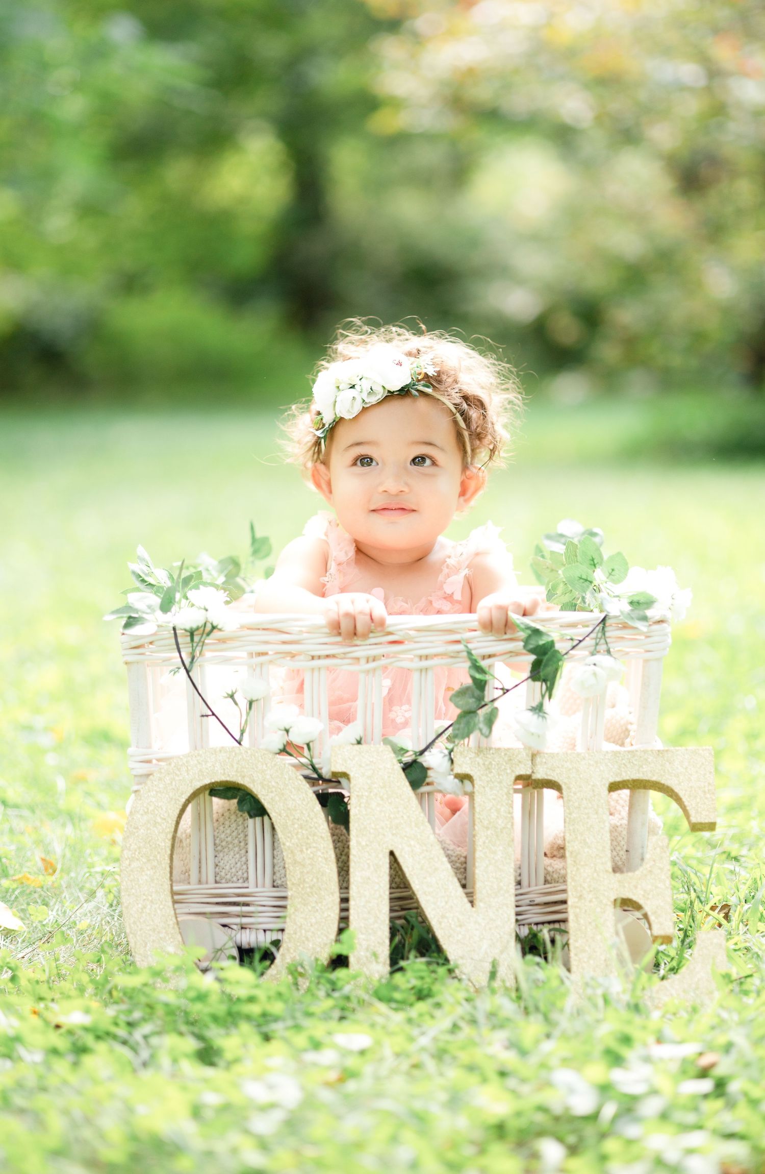 Baby in pink dress poses with decorative ONE sign outdoors among greenery and flowers.