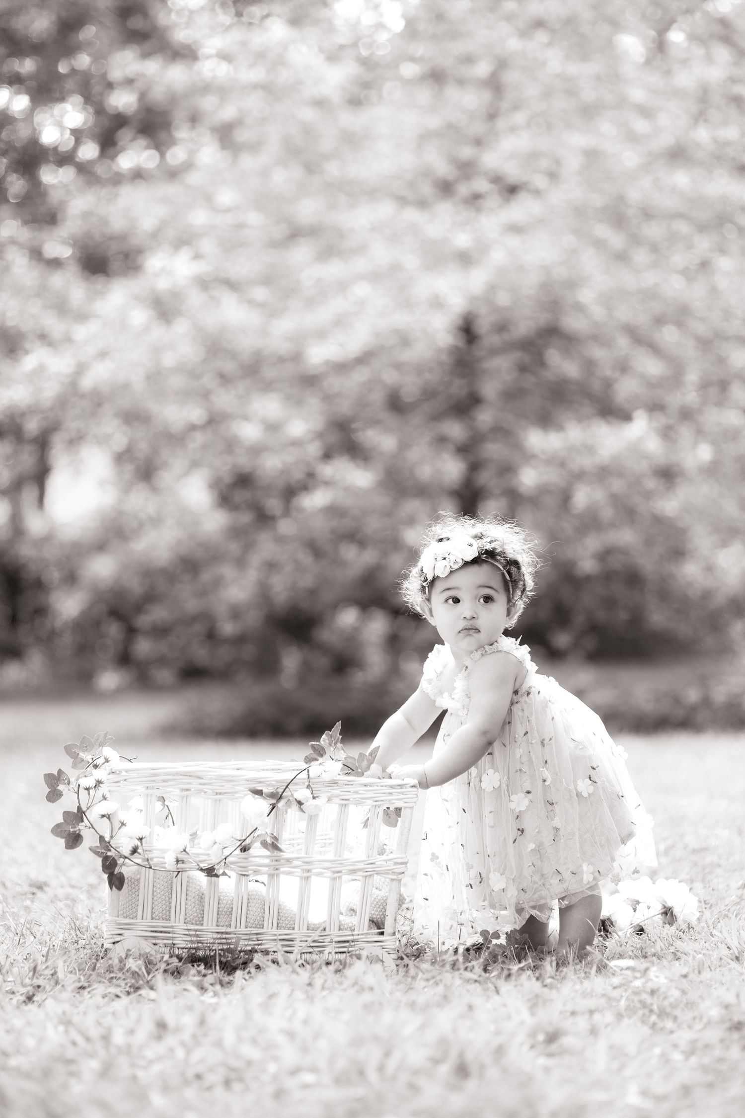 Black and white photo of baby in dress leaning against decorative ONE sign in garden setting.