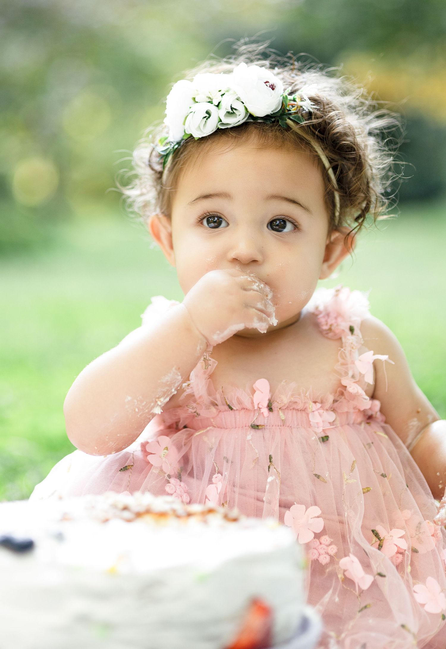 Baby wearing floral crown and pink dress tastes birthday cake during outdoor photoshoot.