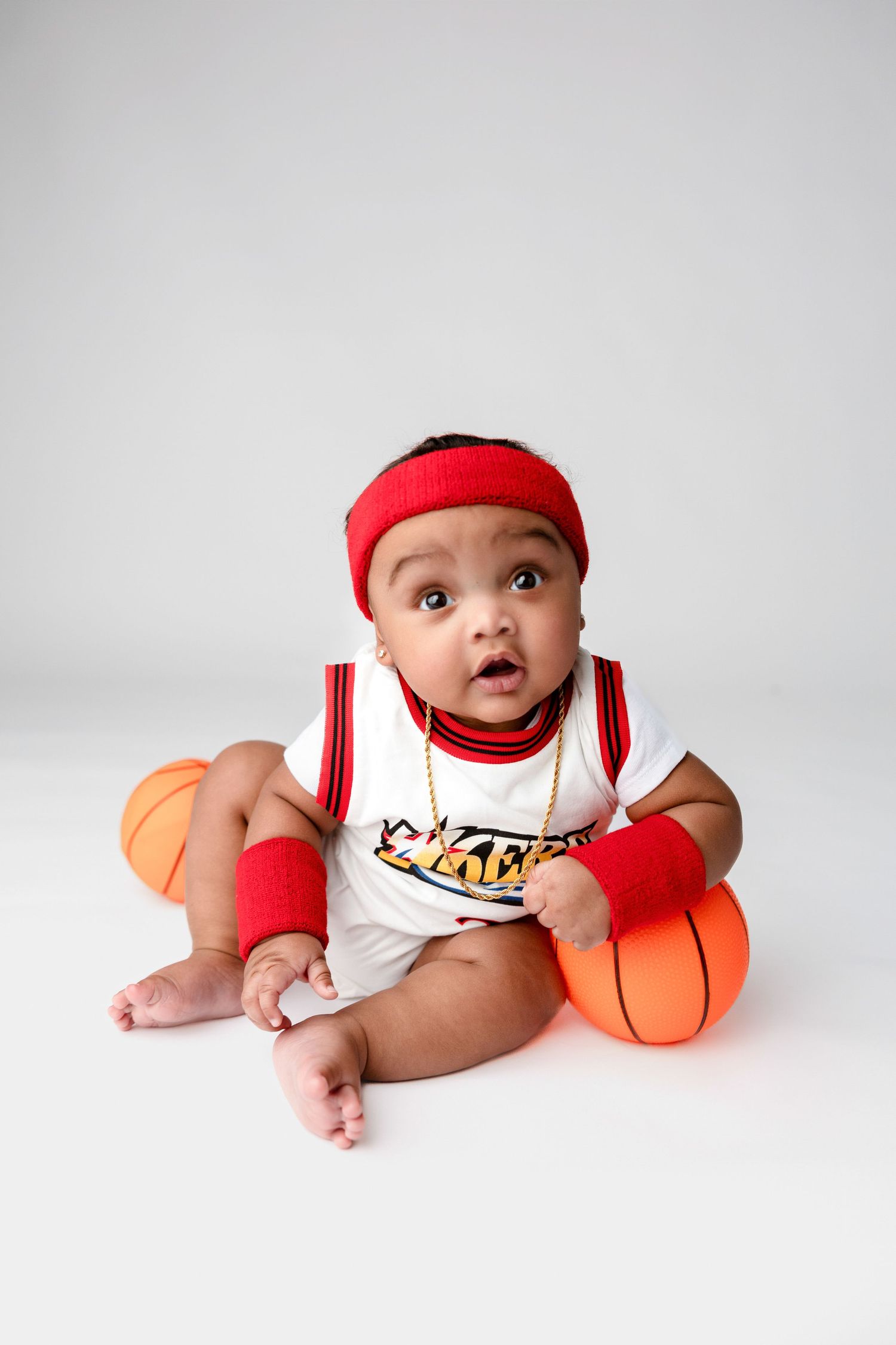 Baby in basketball outfit poses with orange basketballs against white background.