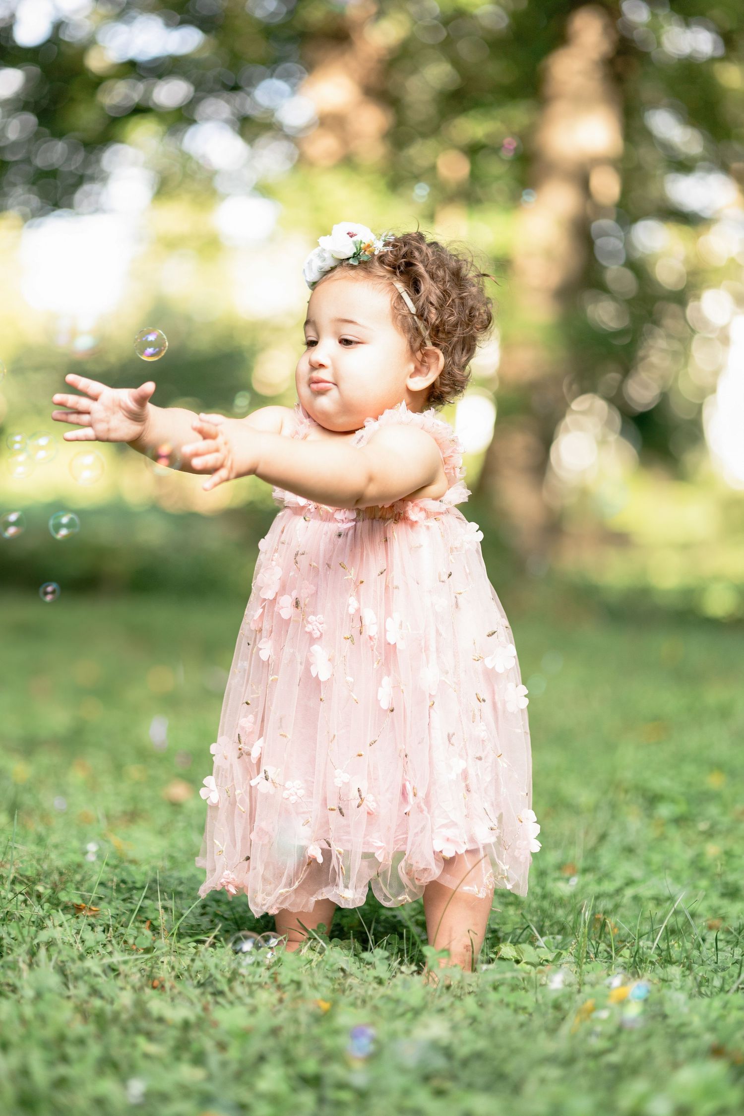 Baby in pink dress reaches for bubbles during outdoor birthday photoshoot.