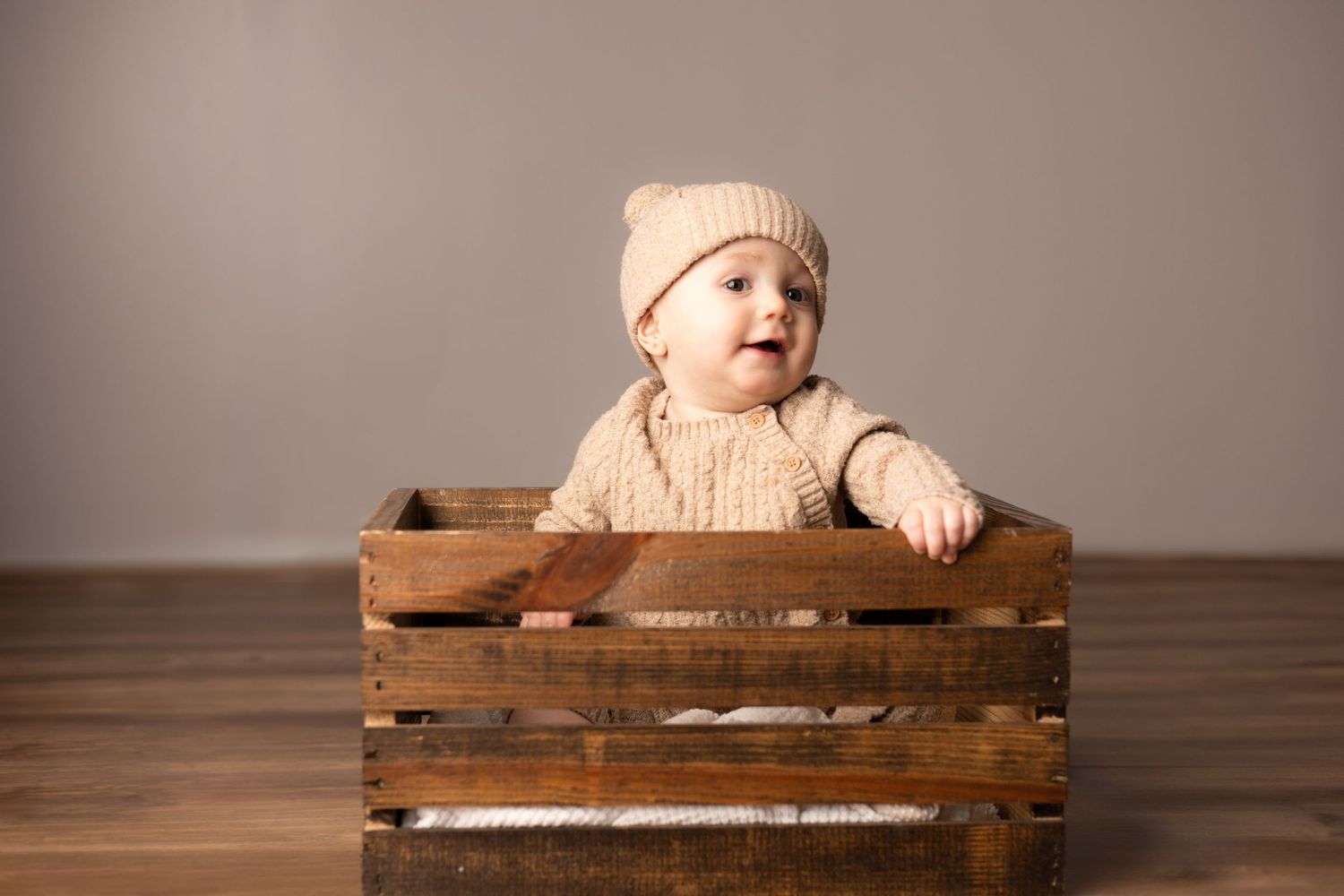 Baby wearing beige knit sweater and hat sits in wooden crate.