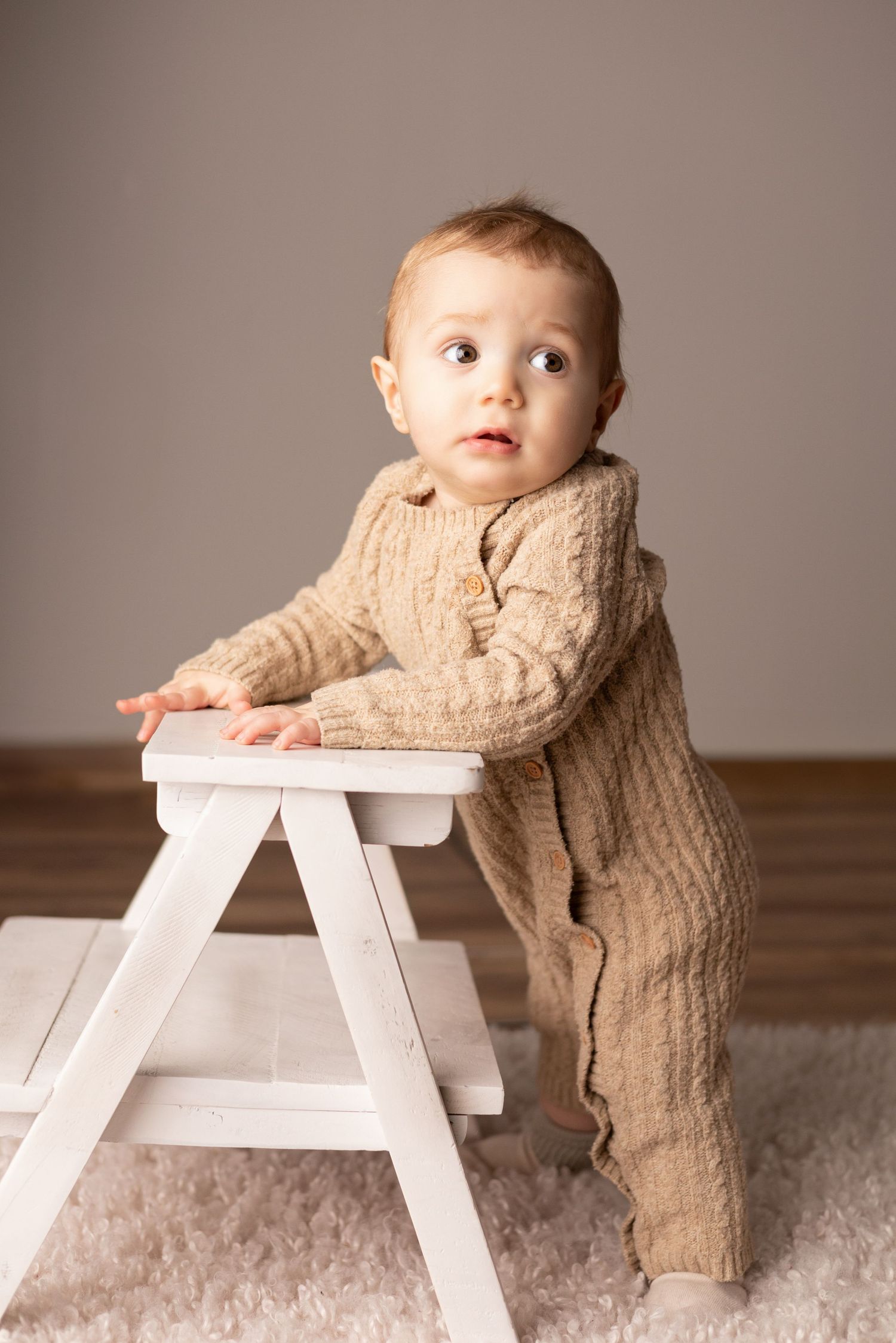 Baby in beige knit outfit standing at white step stool.