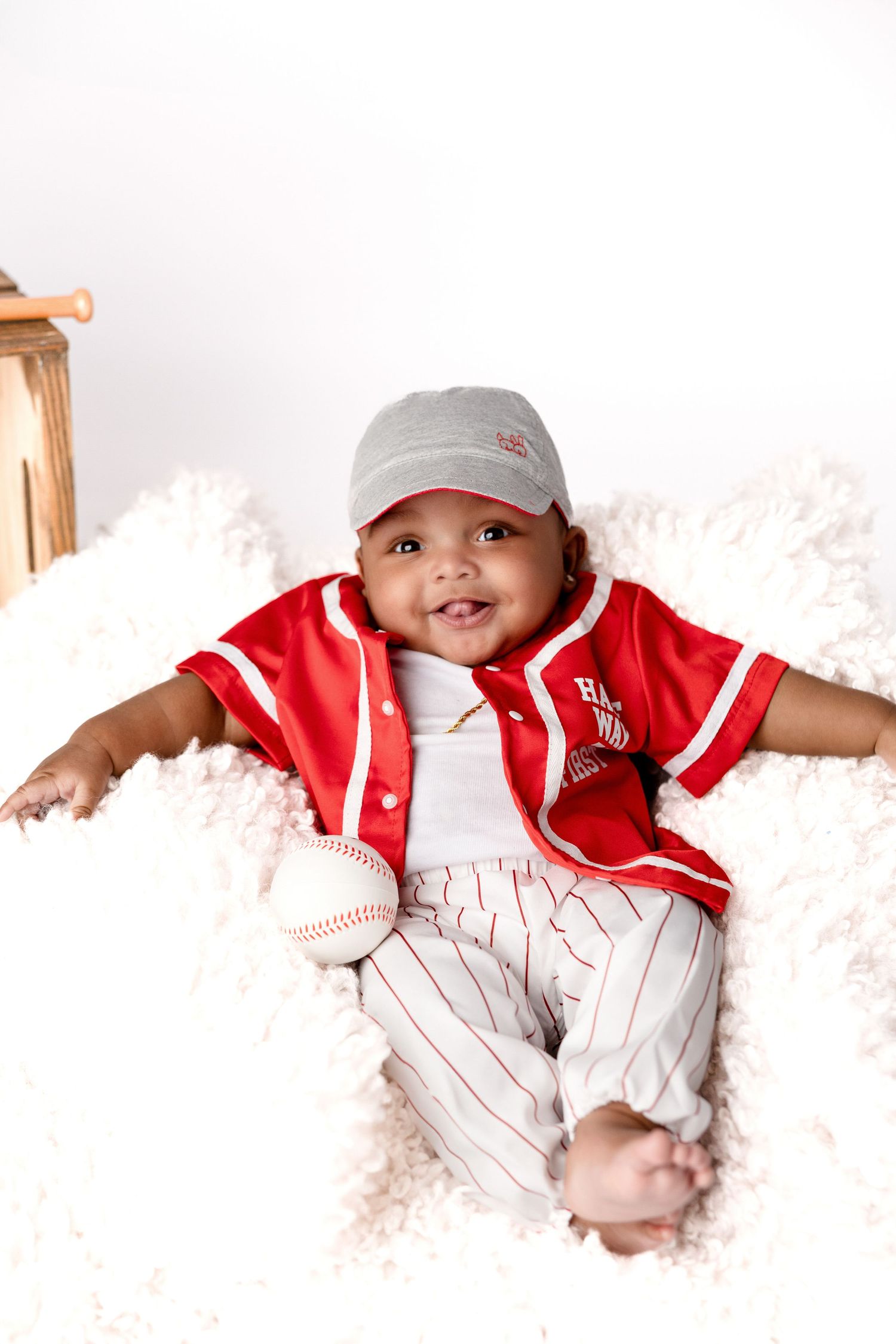 Baby wearing baseball uniform lies on white fluffy blanket with baseball props.