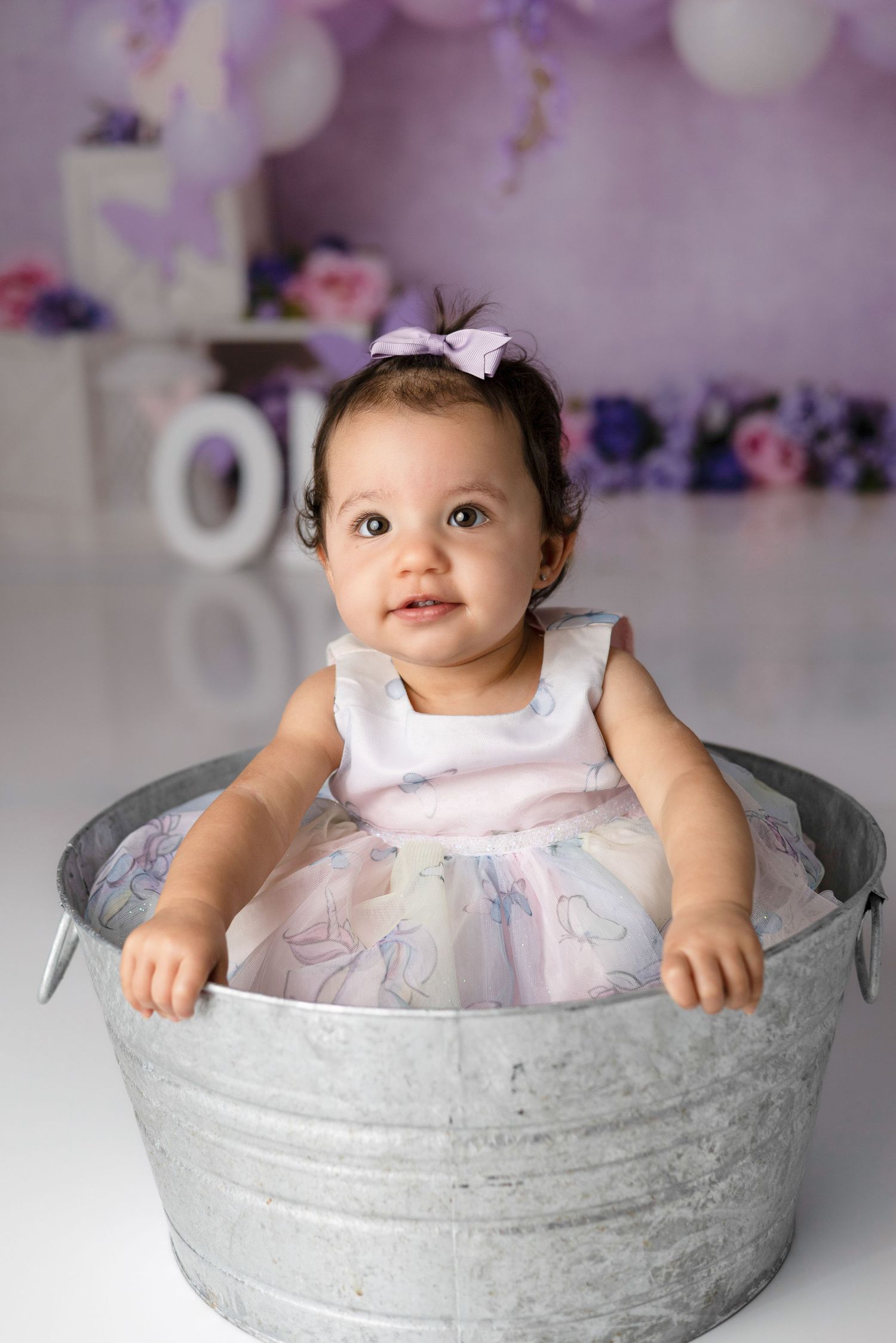 Baby in white dress sits in galvanized metal tub with purple and pink floral backdrop for first birthday photoshoot.
