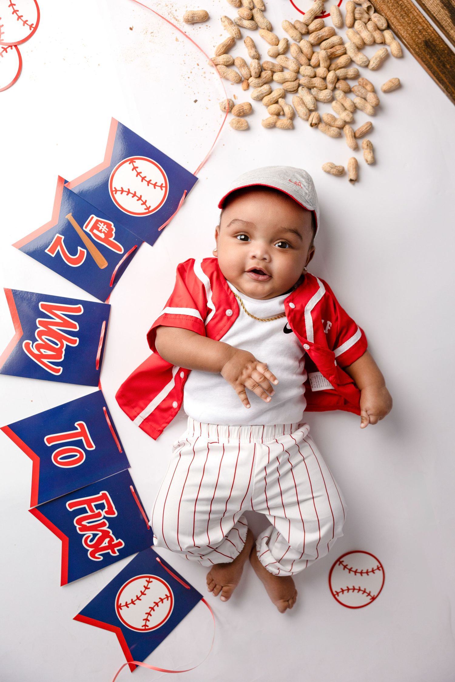 Baby wearing baseball uniform lies on white backdrop with peanuts and baseball decorations.