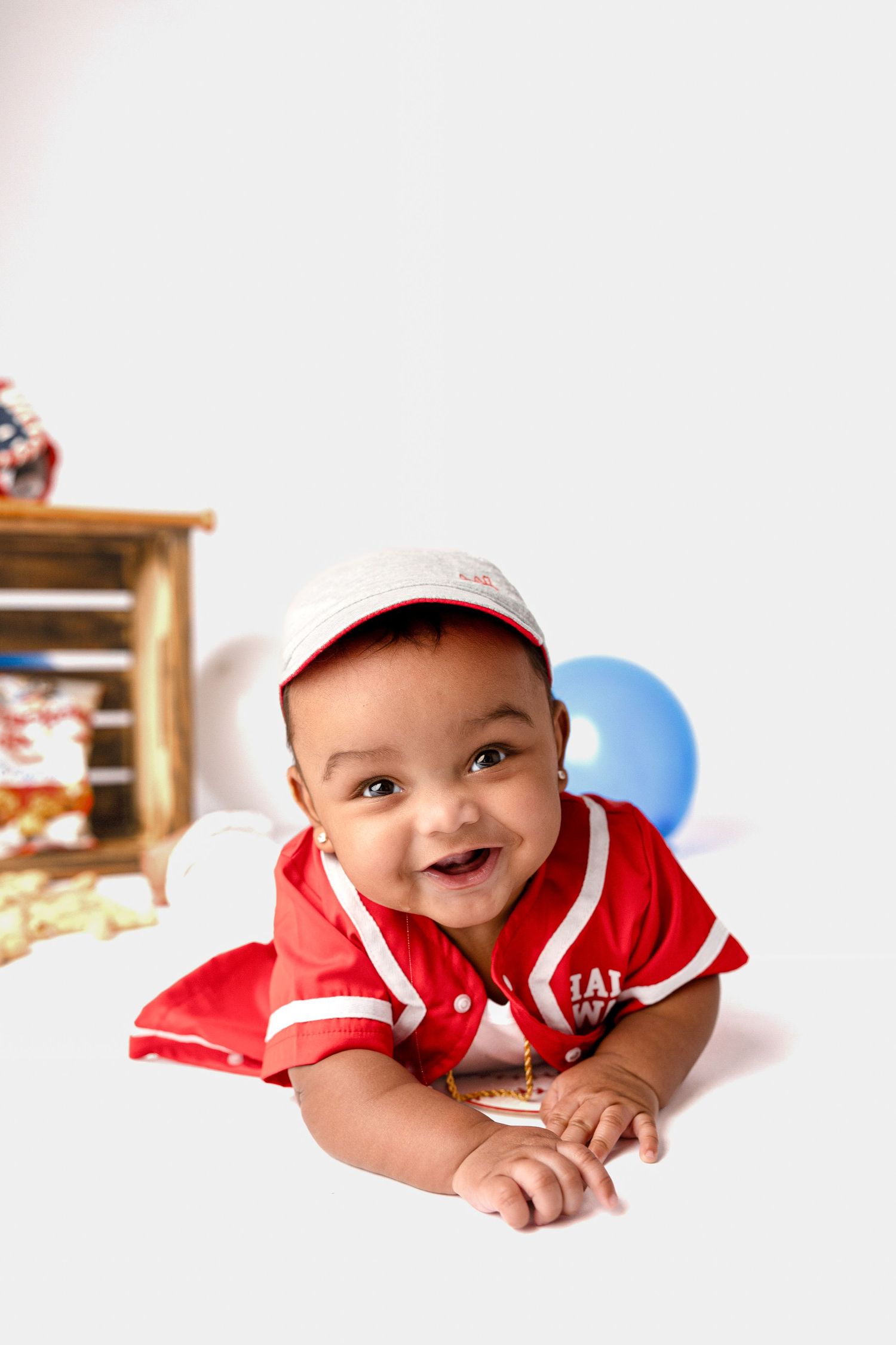Baby in red baseball outfit smiles while lying on white background.