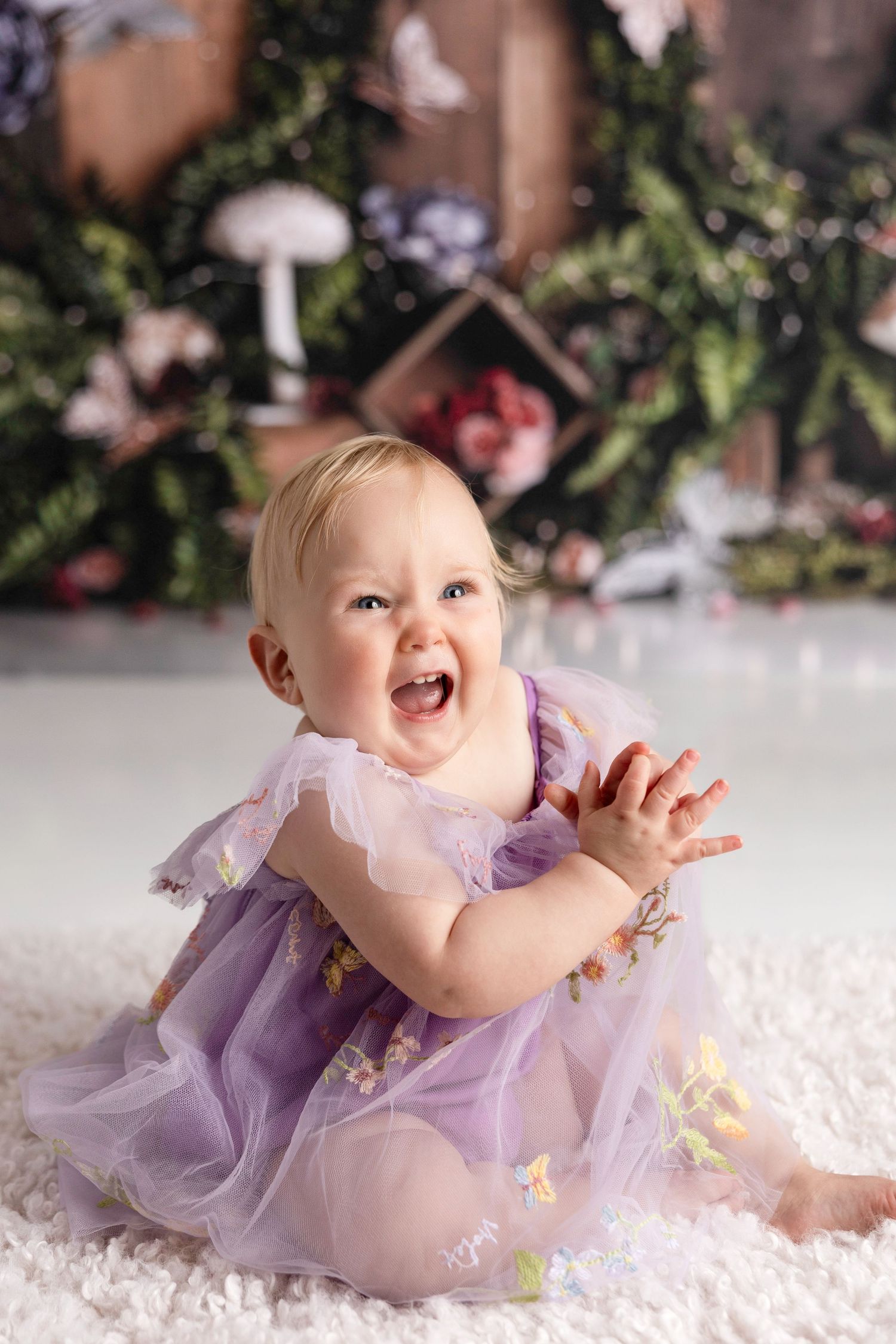 A baby in a purple tulle dress laughs while lying on white carpet during a photo shoot.