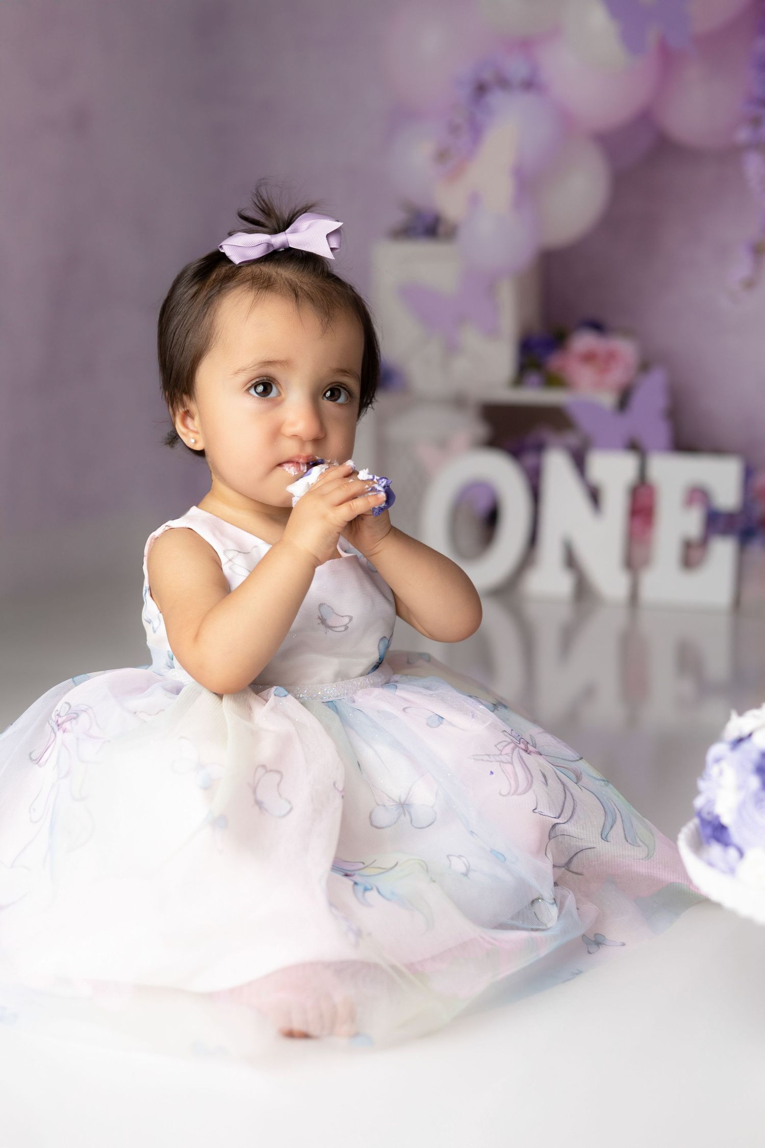Baby in white tulle dress poses with ONE sign and purple decorations for first birthday celebration.