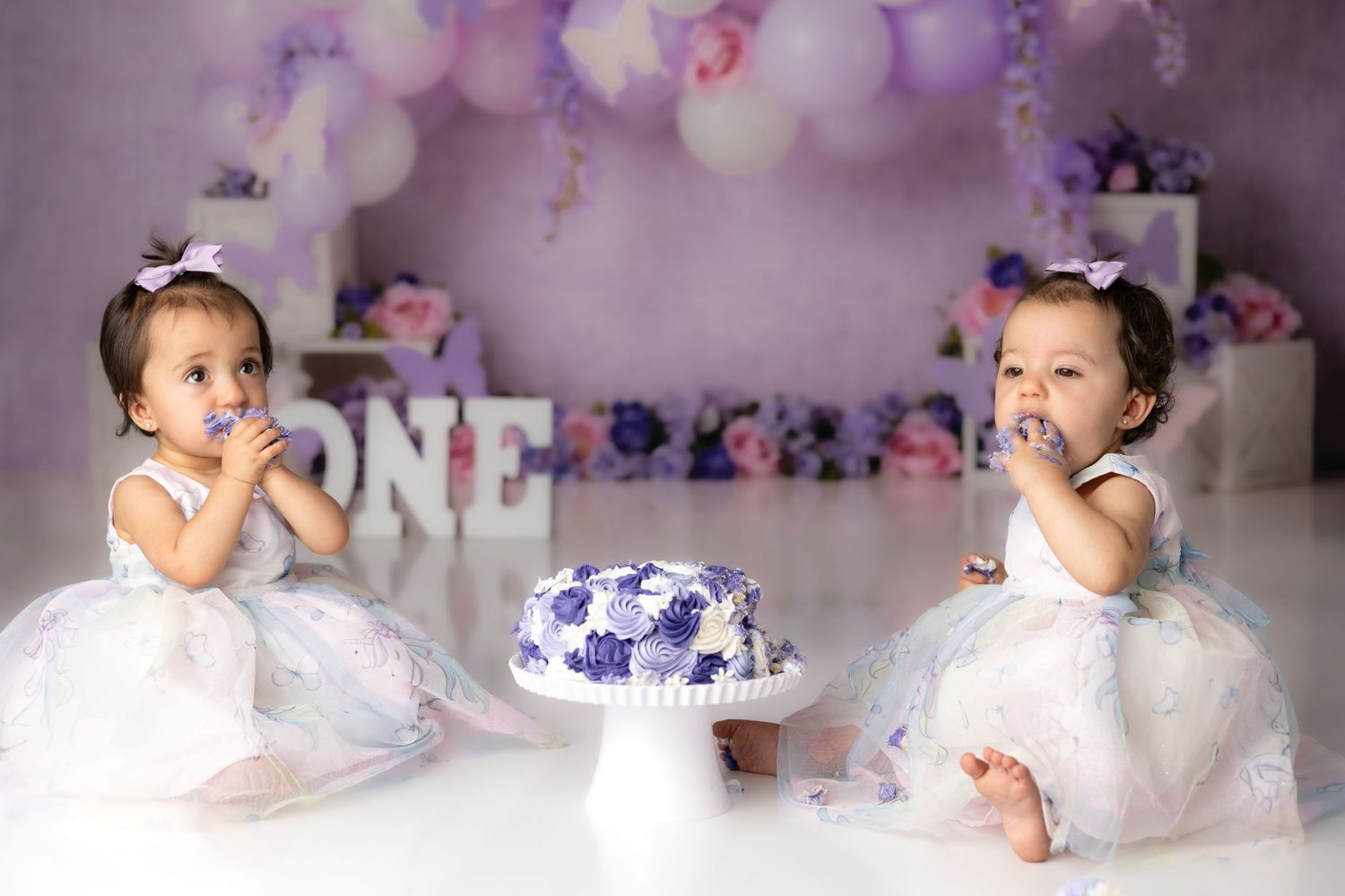 Twin babies in matching white dresses sit beside purple and white birthday cake.