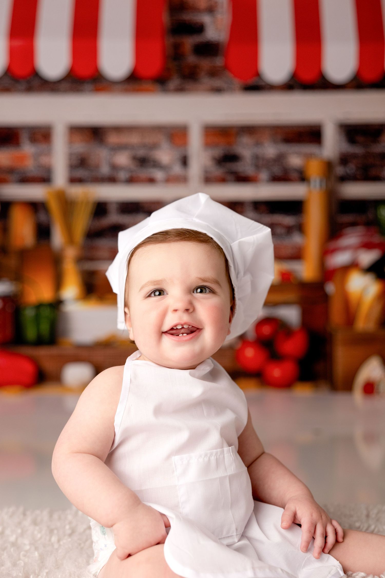 Baby wearing white chef hat smiles against red and white striped background.