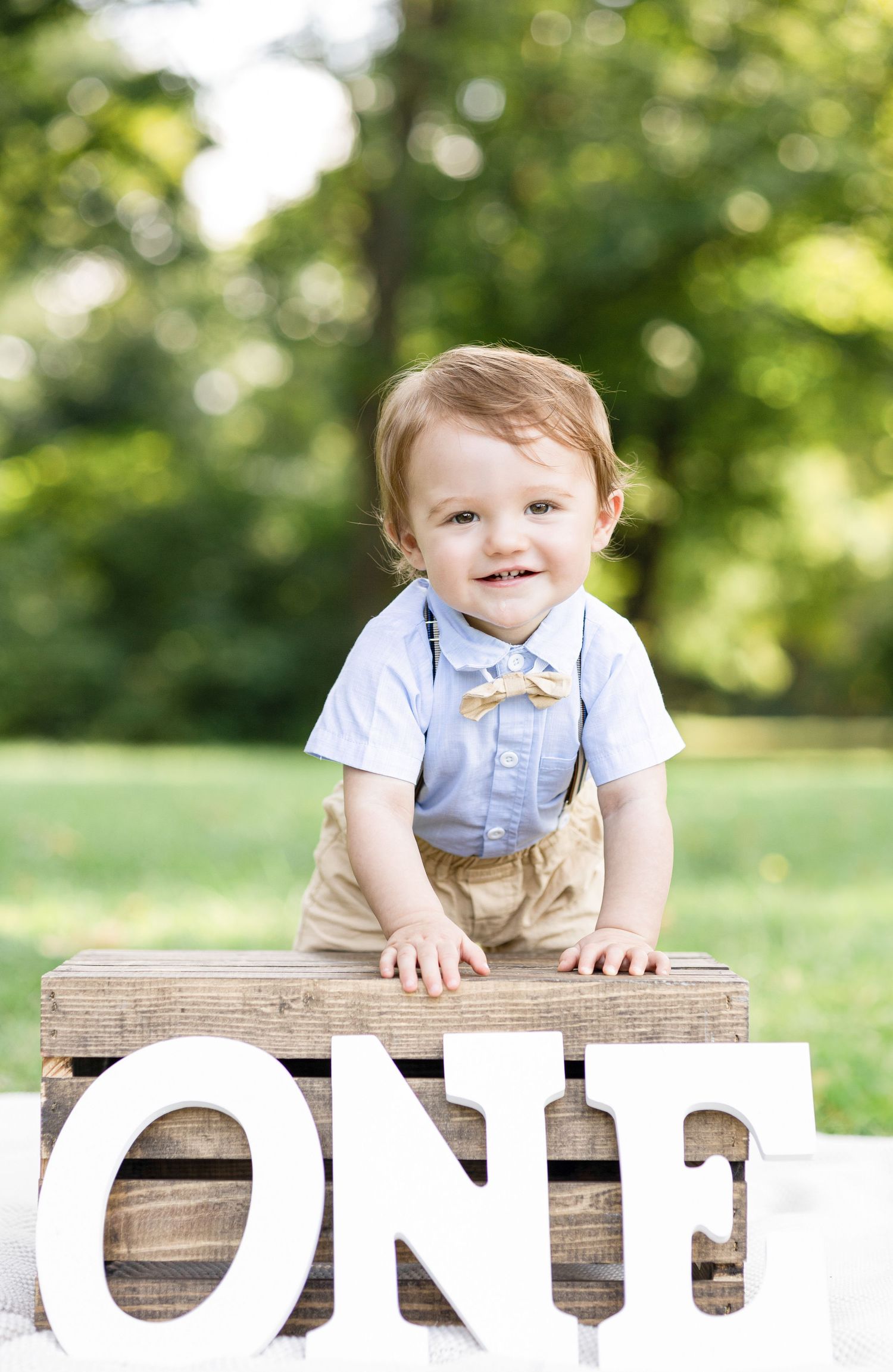 Baby wearing blue bow tie and suspenders poses on wooden blocks outdoors.