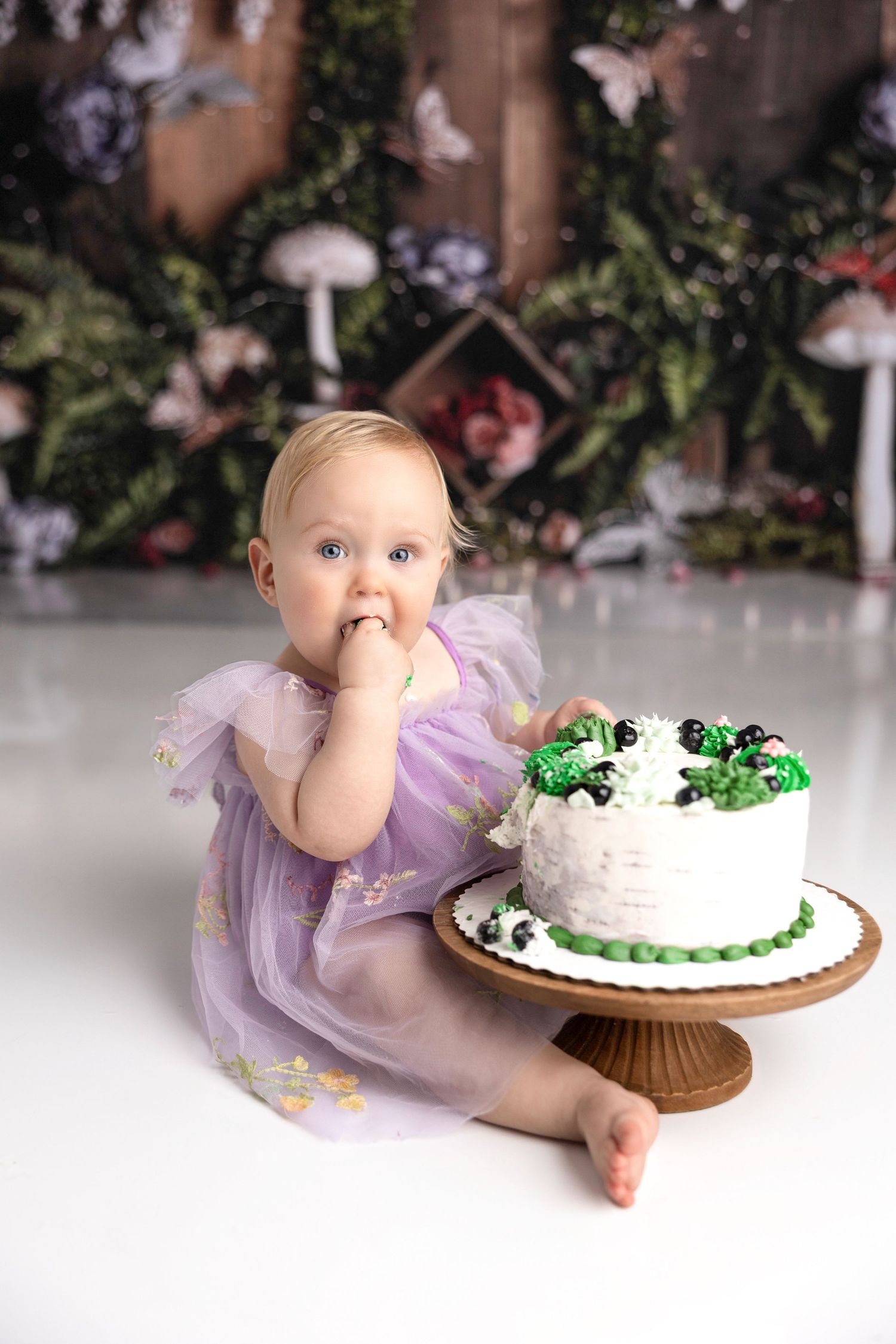 A baby in purple dress reaches for a white birthday cake with green decorations.