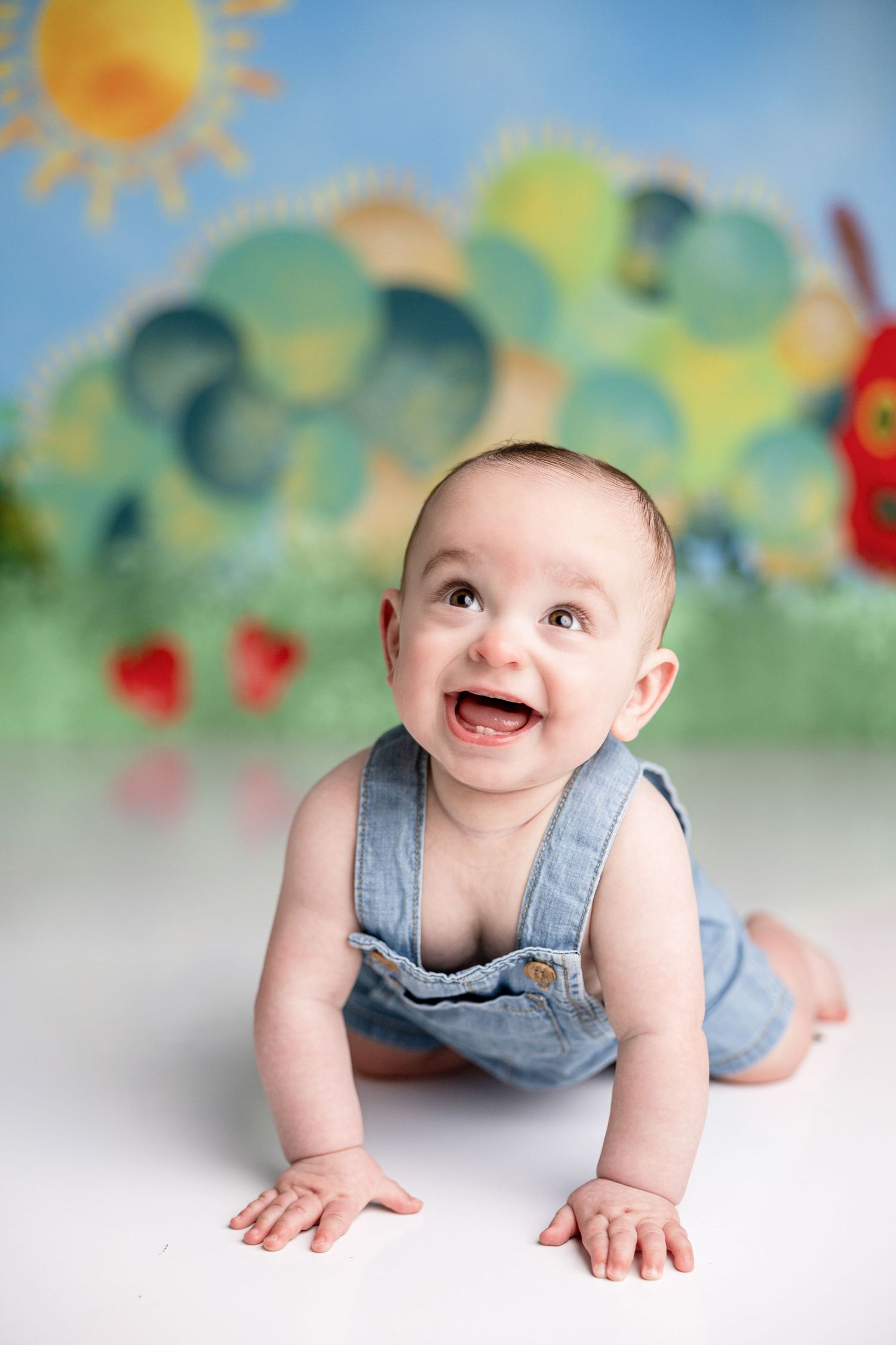 Baby in denim overalls crawling on white floor with painted storybook backdrop.