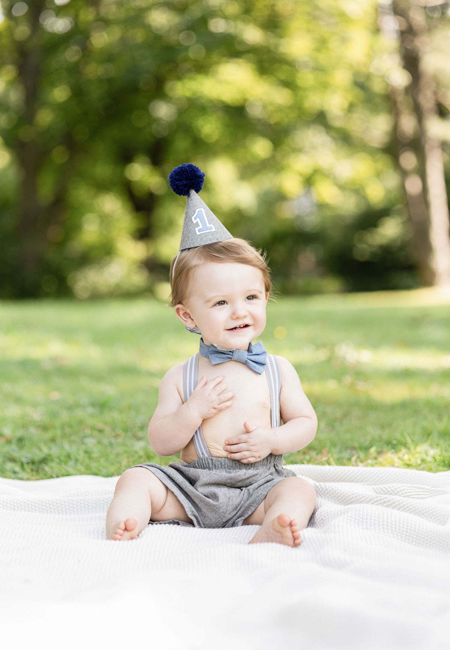 Baby in grey romper and party hat sitting on white blanket in garden setting.