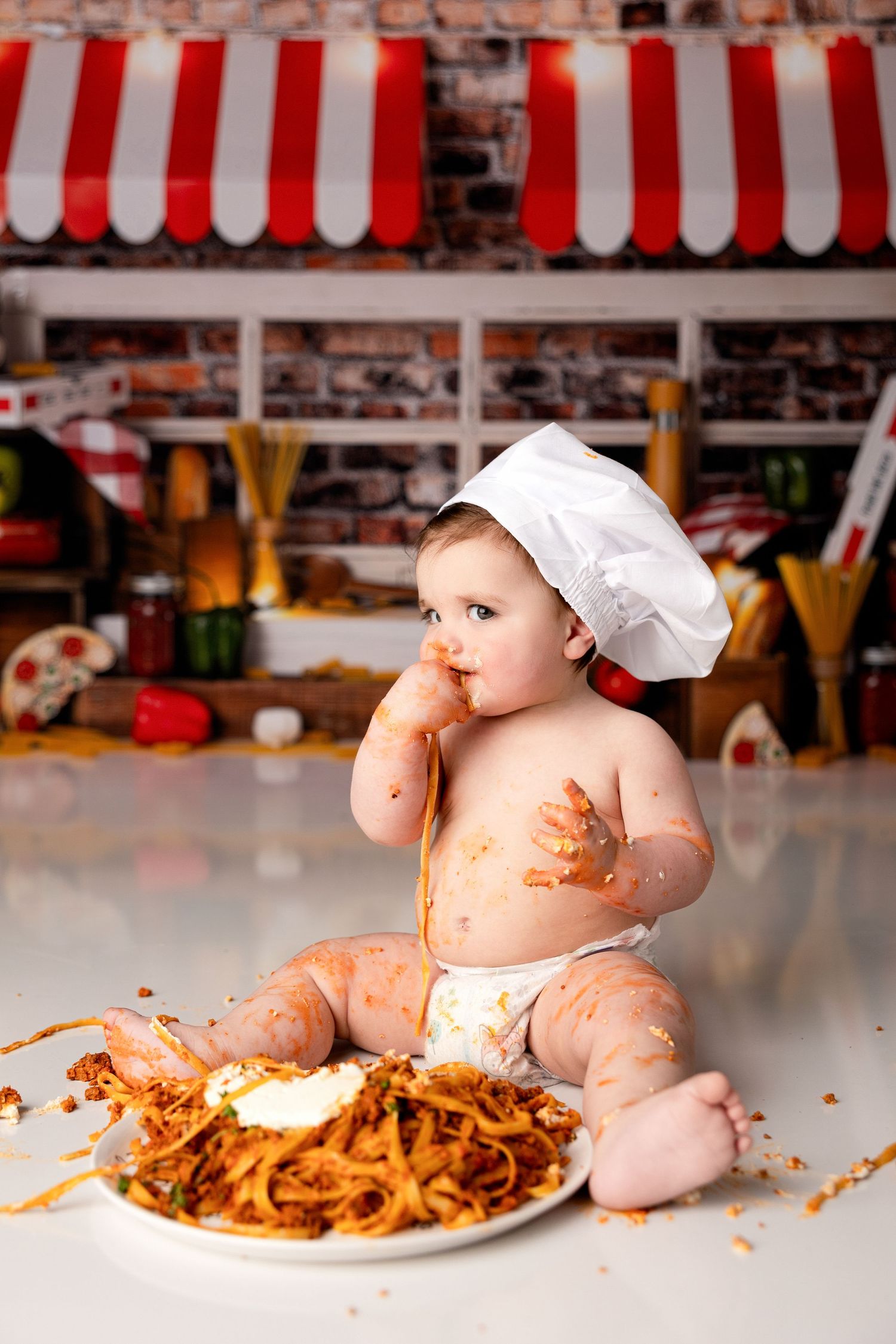 Baby in chef outfit gets messy while eating spaghetti against restaurant backdrop.