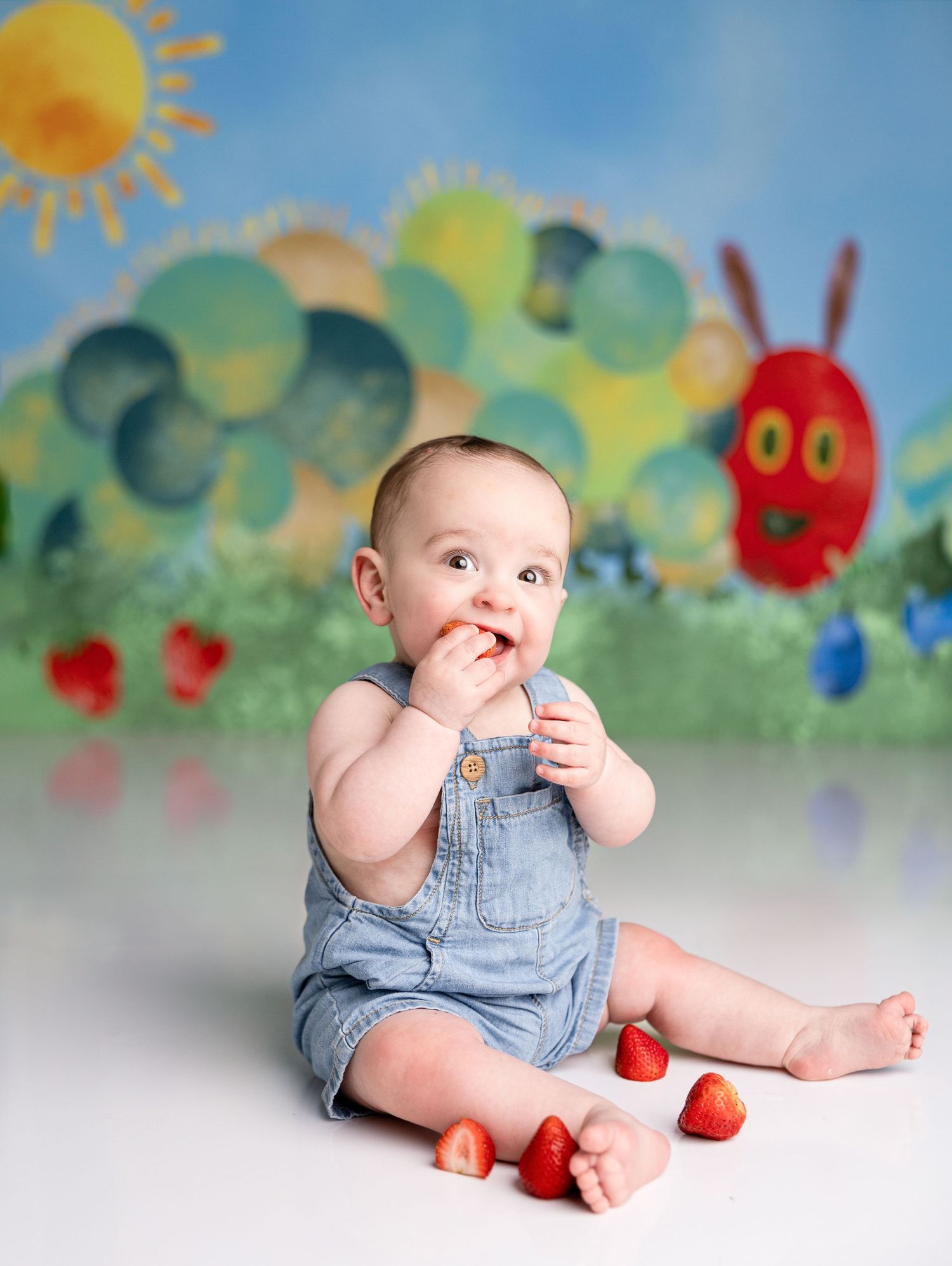 Baby in denim overalls sits with strawberries against painted garden backdrop.