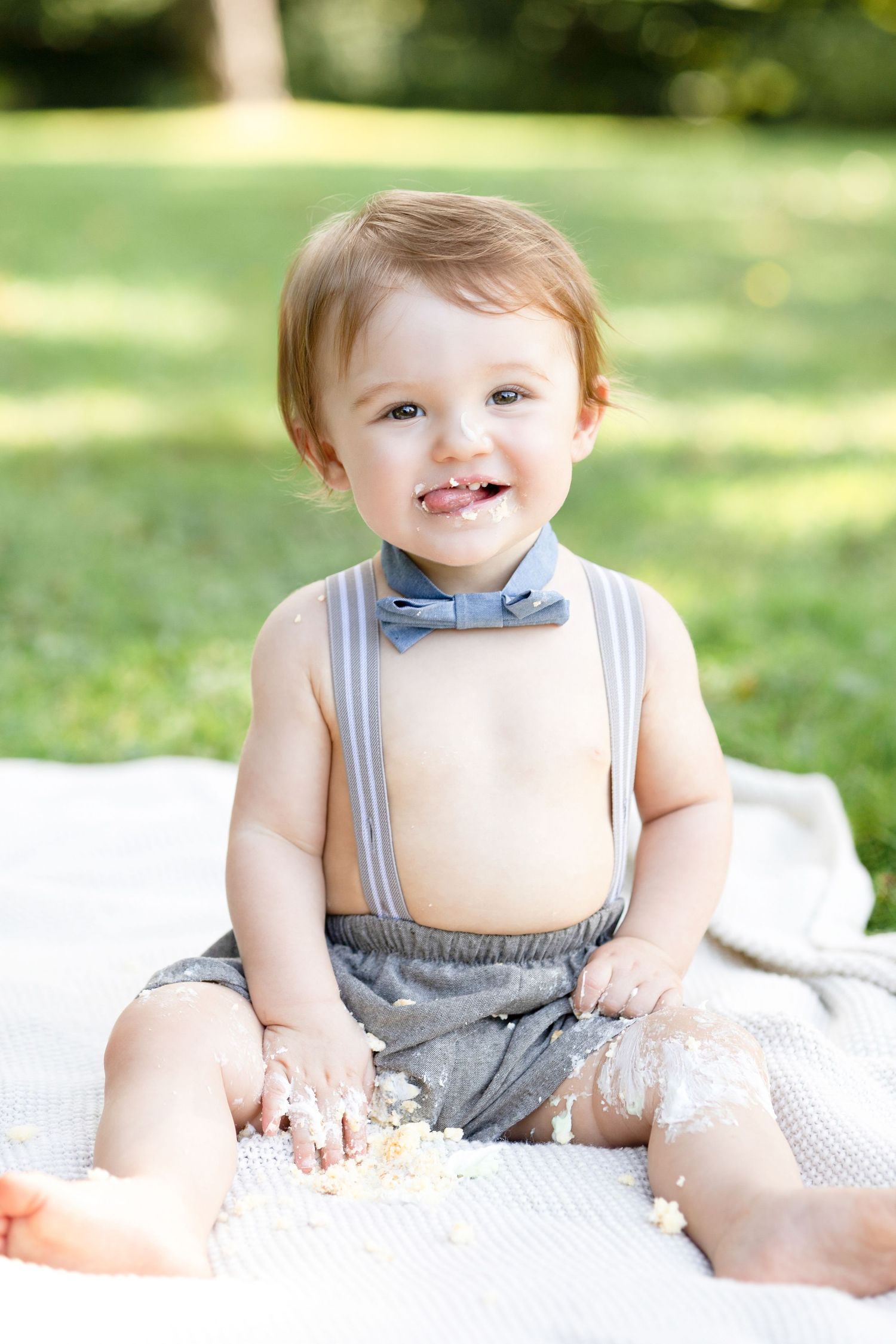 Baby in grey suspender outfit smiling while sitting on white blanket outdoors.