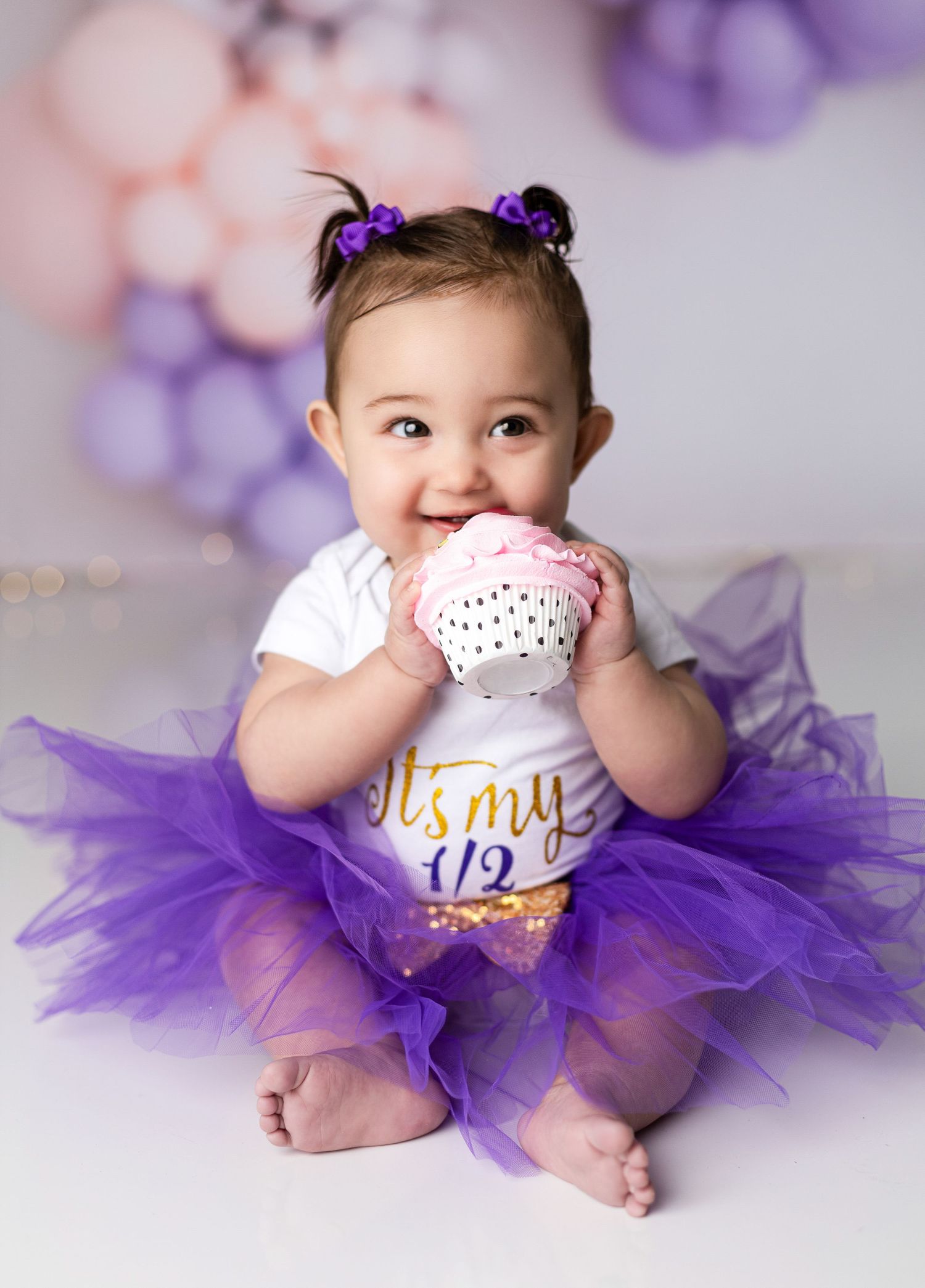 Baby in purple tutu and white onesie sitting against purple balloon backdrop.