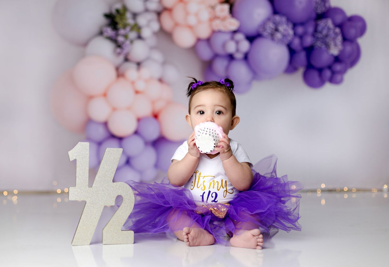 Baby in purple tutu dress with milestone numbers and balloons in background.