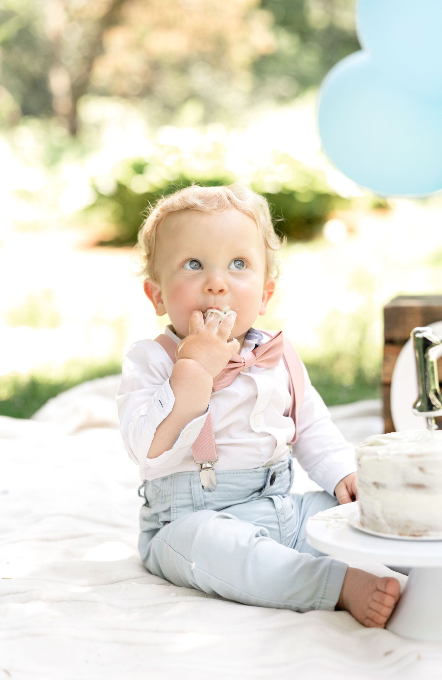 A baby in white and gray outfit sits on white blanket with blue balloons in background.