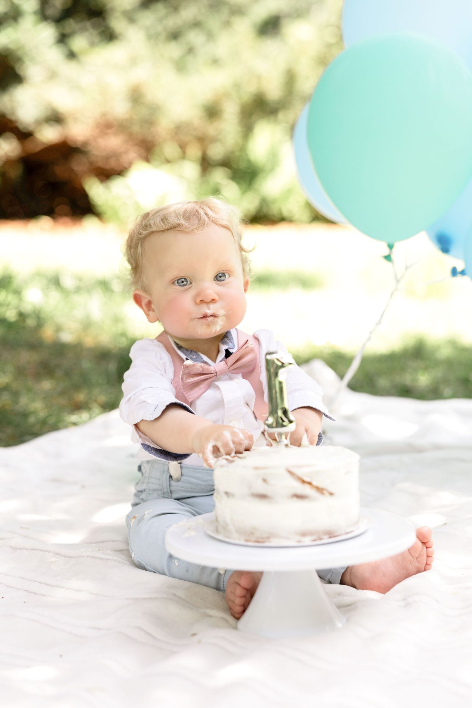 A baby in formal attire explores their first birthday cake on a white blanket outdoors.