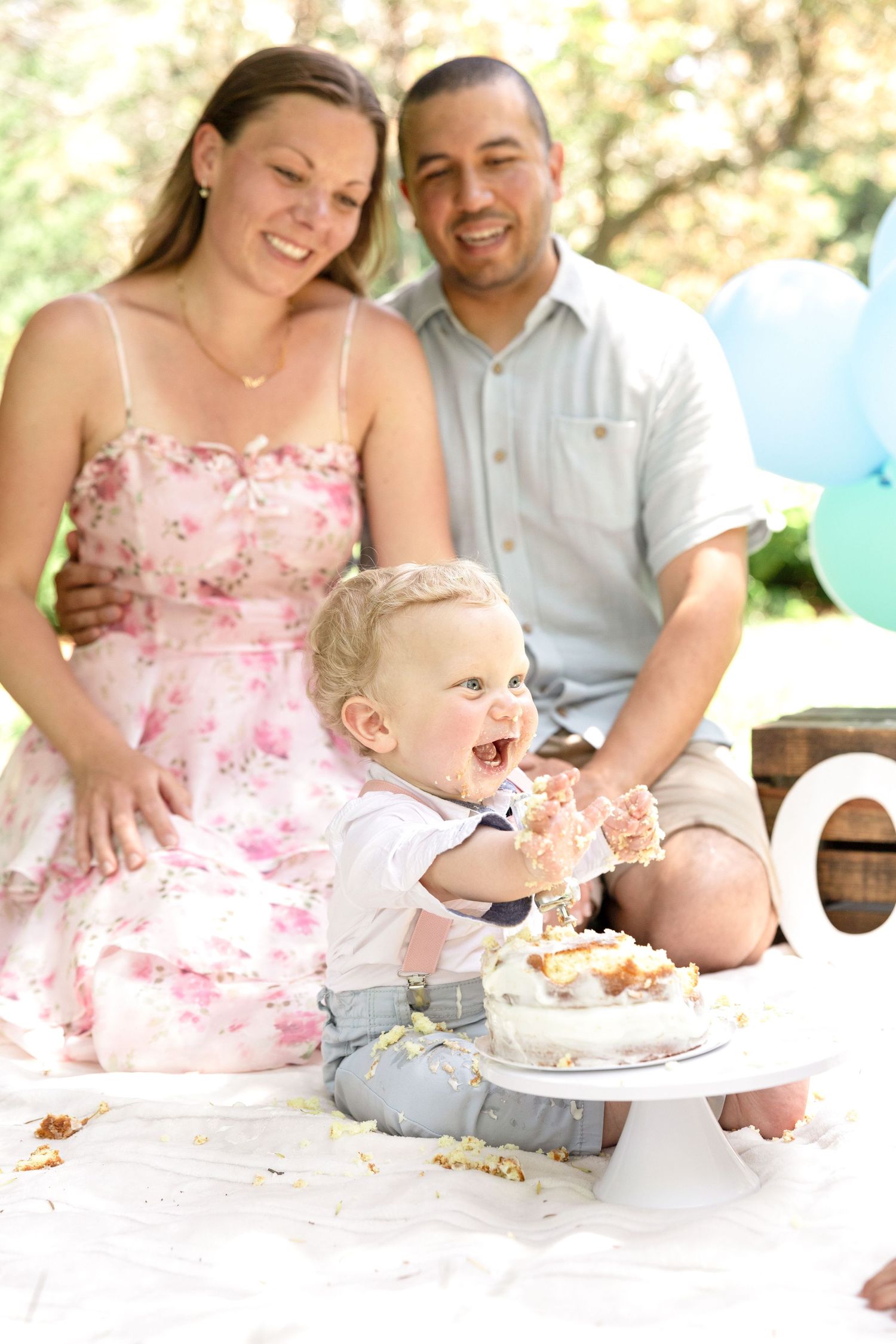 A family celebrates a cake smash photo session outdoors with blue and mint balloons.