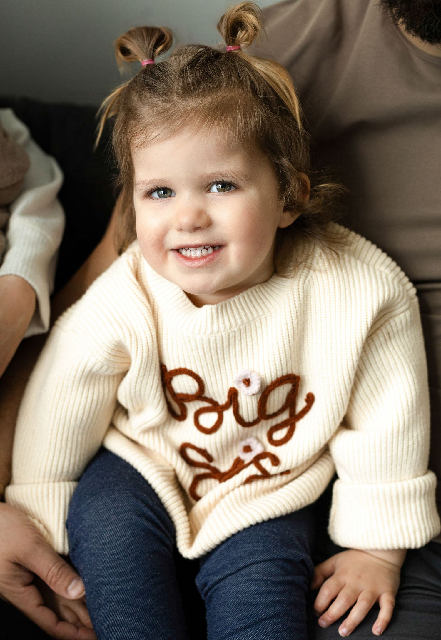 Toddler wearing cream sweater and navy pants smiles while sitting on couch.