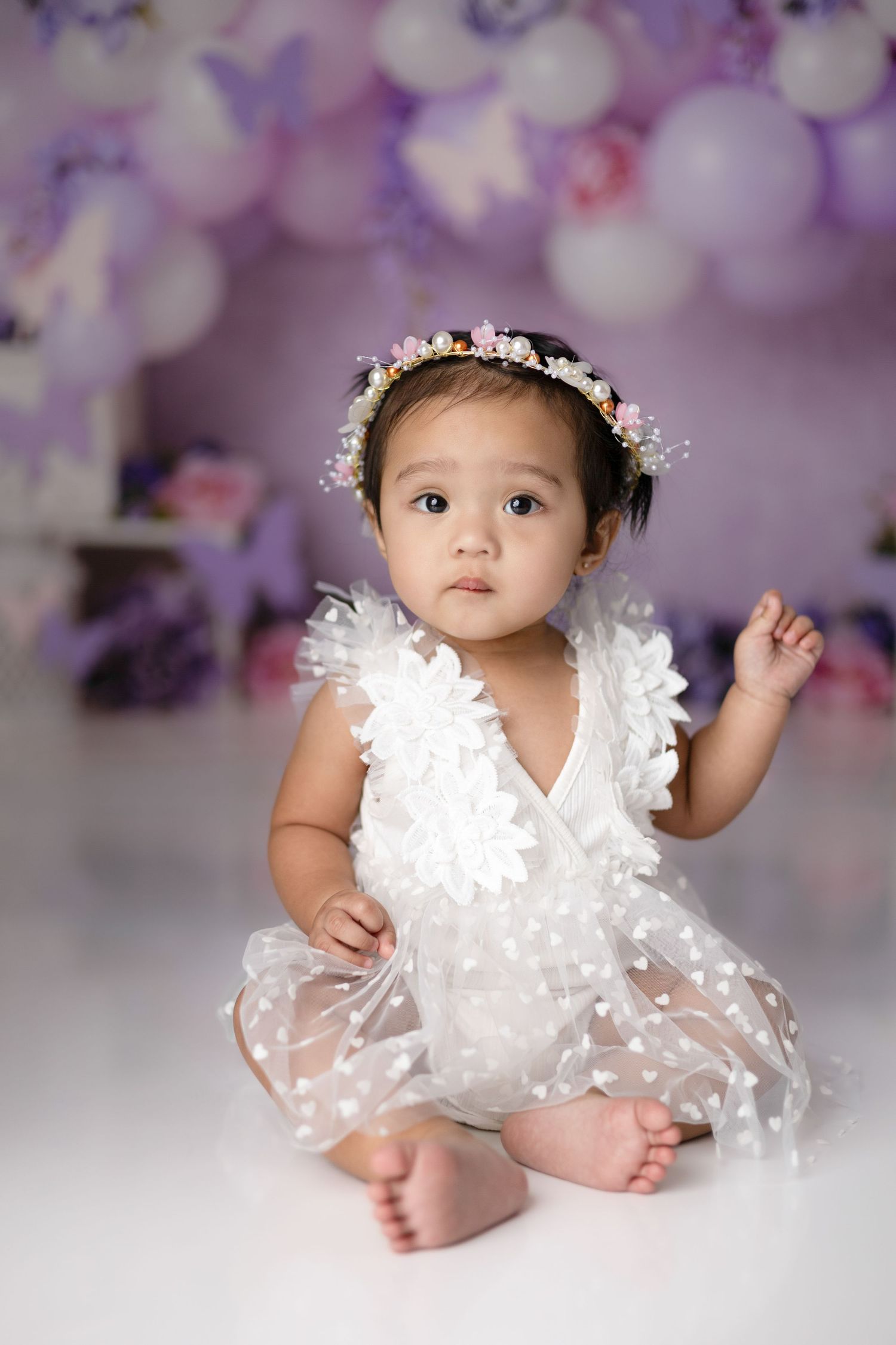 Baby wearing white lace dress and floral crown against purple bokeh background.