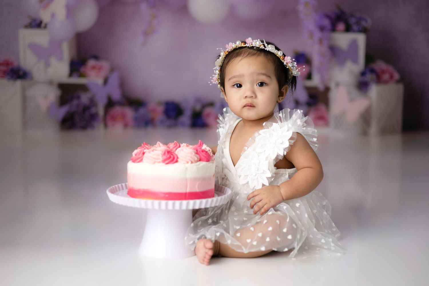 Baby in white dress reaching toward pink birthday cake during photo session.