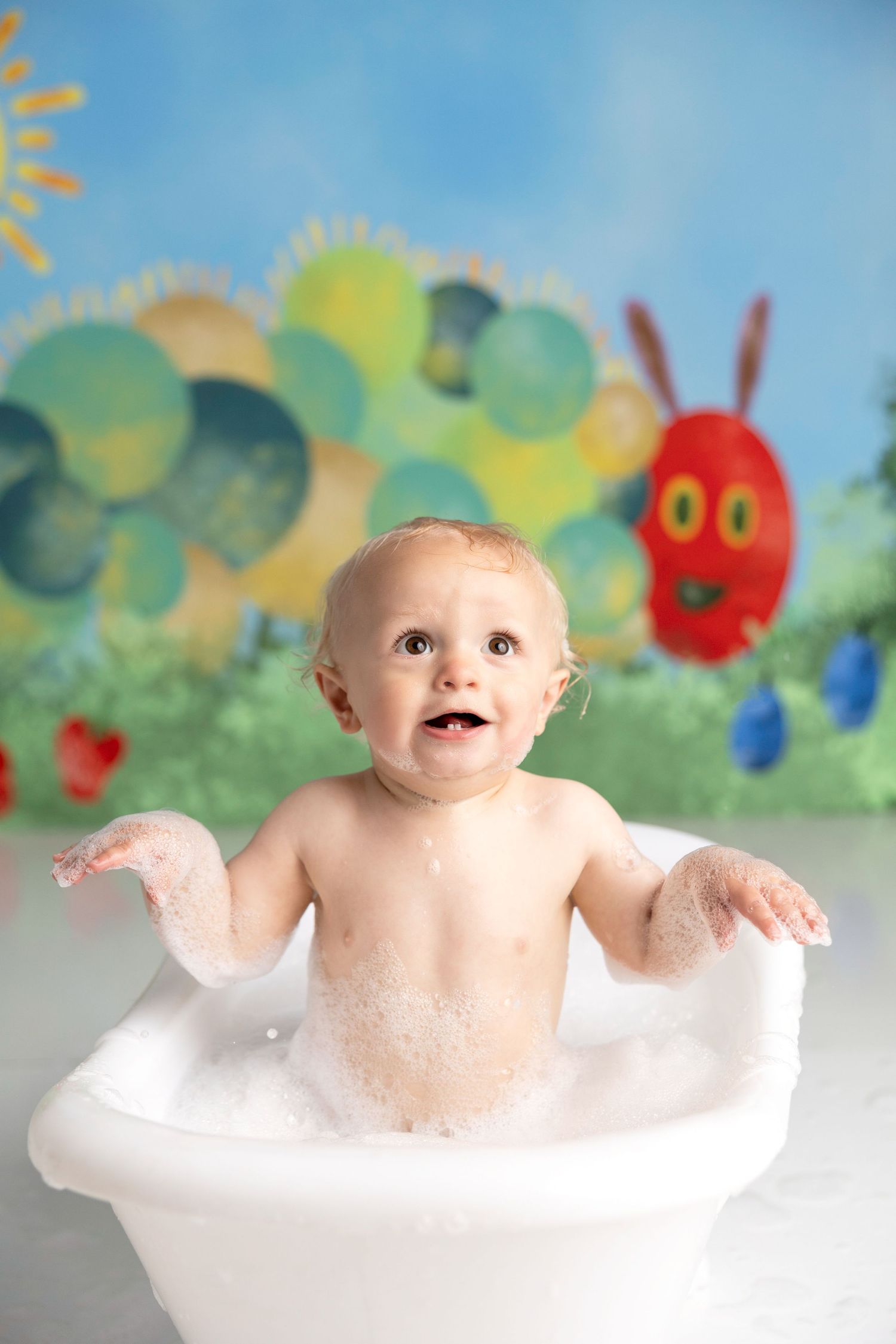 Baby splashing in bubble bath with colorful wall art featuring The Very Hungry Caterpillar theme in background.