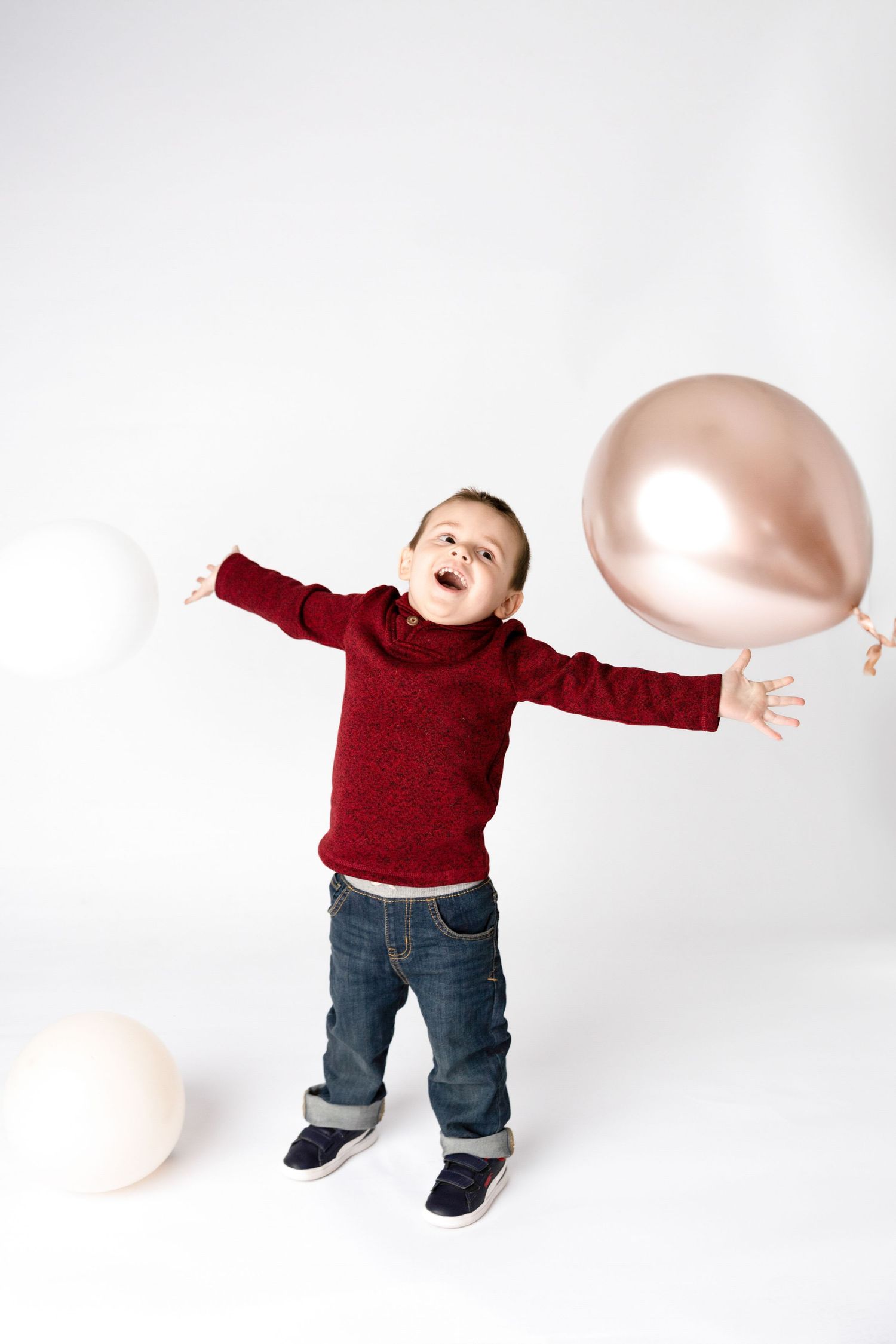 Excited child in red sweater playing with pink and white balloons against white background.