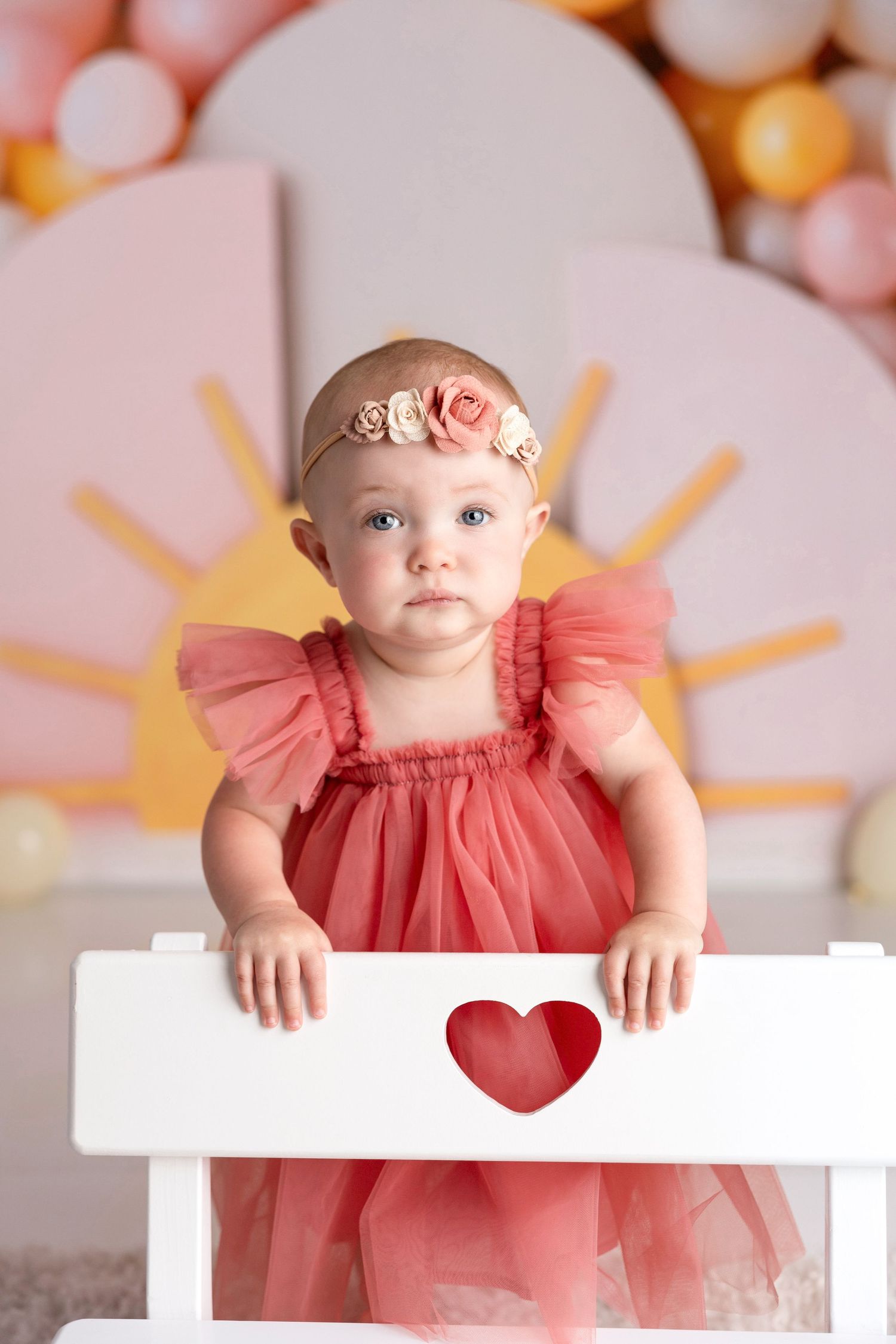 A baby in coral tulle dress poses against a pink balloon backdrop with sun decorations.