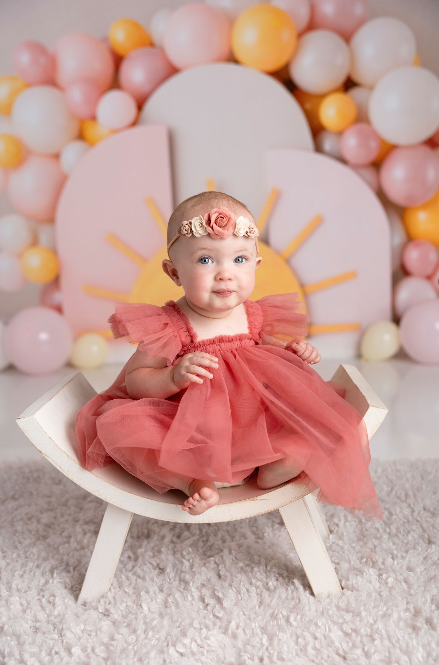 A baby wearing a coral dress sits on a white bench with balloon decorations.