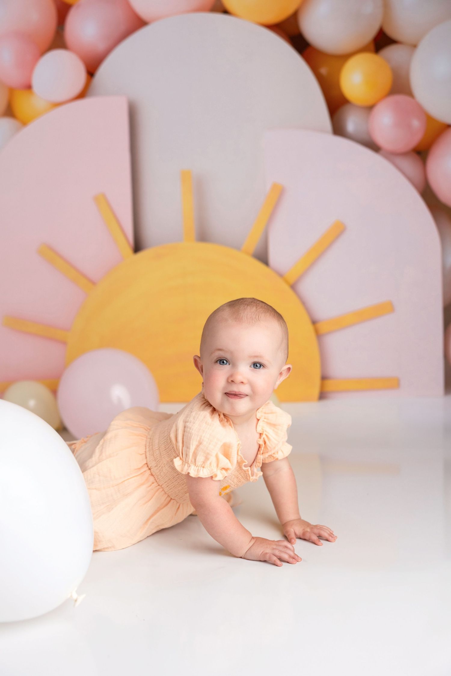 A baby in peach outfit crawls on white floor with sun and balloon decorations behind.